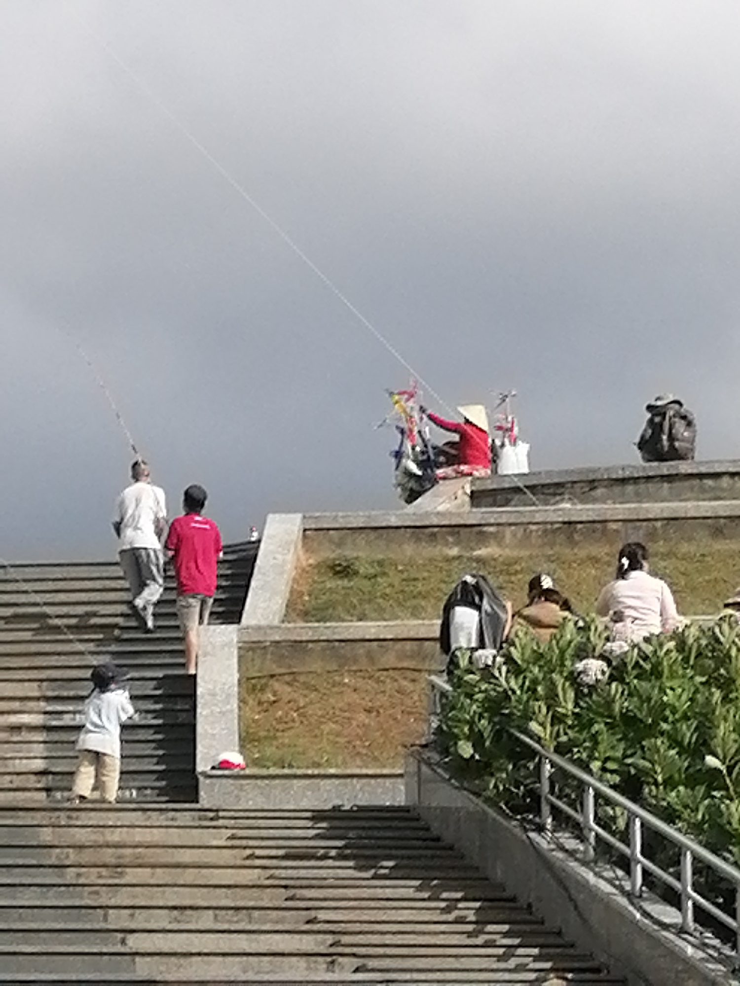 The image depicts an outdoor scene with a cloudy sky. In the foreground, a child in a light-colored shirt and tan pants stands on stone stairs. Further up the stairs, two men are walking. The man in front is wearing a white shirt and gray pants, carrying a fishing rod. The man behind him wears a red shirt and shorts. Above them, a woman in a conical hat and red top is seated, with various items displayed around her. To the right, a person sits near a backpack, and further down, two individuals are seated in front of lush green bushes. A metal railing runs along a pathway beside the bushes. The architecture suggests a public space with tiered levels and stone construction, possibly a park or a monument. The presence of fishing rods and the overall atmosphere suggests a leisurely activity. Given the context of Da Lat, Vietnam, this scene could be from a location like the Da Lat Flower Gardens or a similar public recreational area.