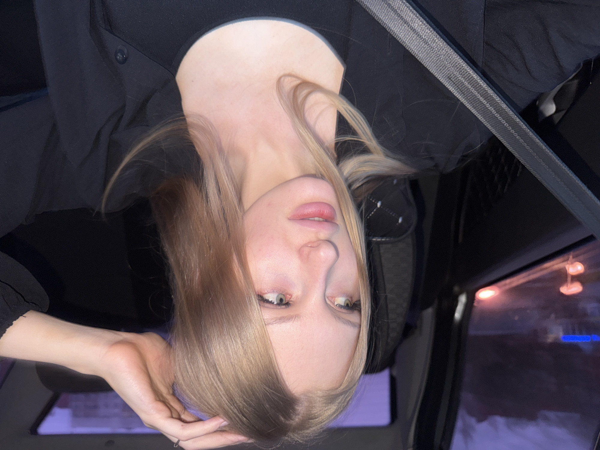 A young woman with fair skin and blonde hair, wearing a black garment, is seated inside a vehicle. She is oriented upside down in the frame. A seatbelt is visible across her upper body. Outside the vehicle's window, blurred lights suggest a nighttime setting in an urban area. The location may be in Perm, Russia, as requested.