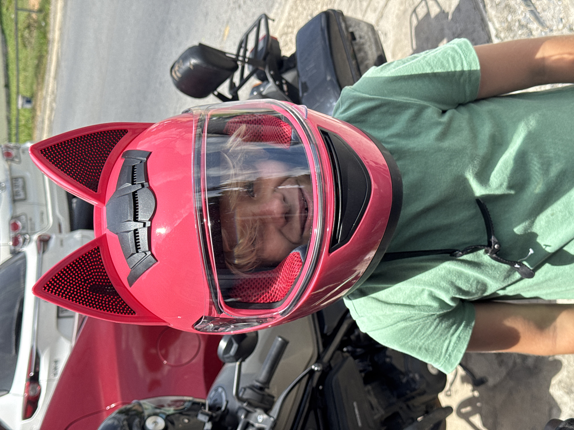 A young person wearing a bright pink full-face helmet with cat ears and a clear visor smiles into the camera. They are wearing a green t-shirt and a black strap is visible around their neck. The helmet has red mesh accents inside the ears and on the cheek pads. Behind the person, a motorcycle is visible, and further in the background, a red car and two white cars are parked. The setting appears to be outdoors on a paved surface, possibly a street or parking lot, with some vegetation and a curb visible. The lighting suggests it is daytime with bright sunlight.