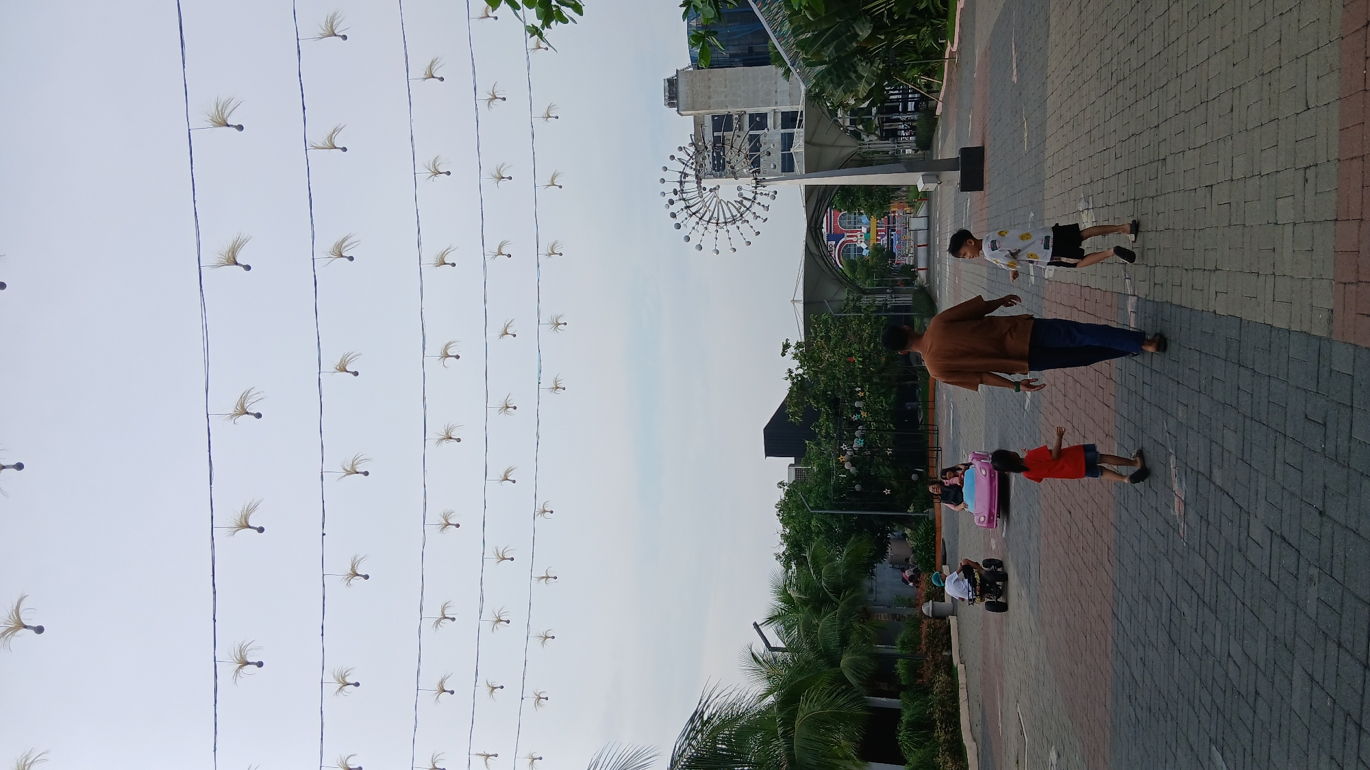 In an outdoor, urban setting in the City of Medan, Indonesia, a man in a brown t-shirt and blue pants walks away from the camera, followed by two young children. To the left, a young girl in an orange t-shirt and blue shorts walks ahead of them. To the right, a young boy in a white t-shirt with a pattern and black shorts walks beside the man, looking back. Further left, a small pink toy car carries two people. In the background, palm trees and other lush greenery line a paved pathway. Overhead, strings of decorative lights with fluffy accents are suspended across the sky. The scene appears to be a public area, possibly a park or promenade, with a large, modern building and a unique kinetic sculpture visible in the distance. The sky is overcast.