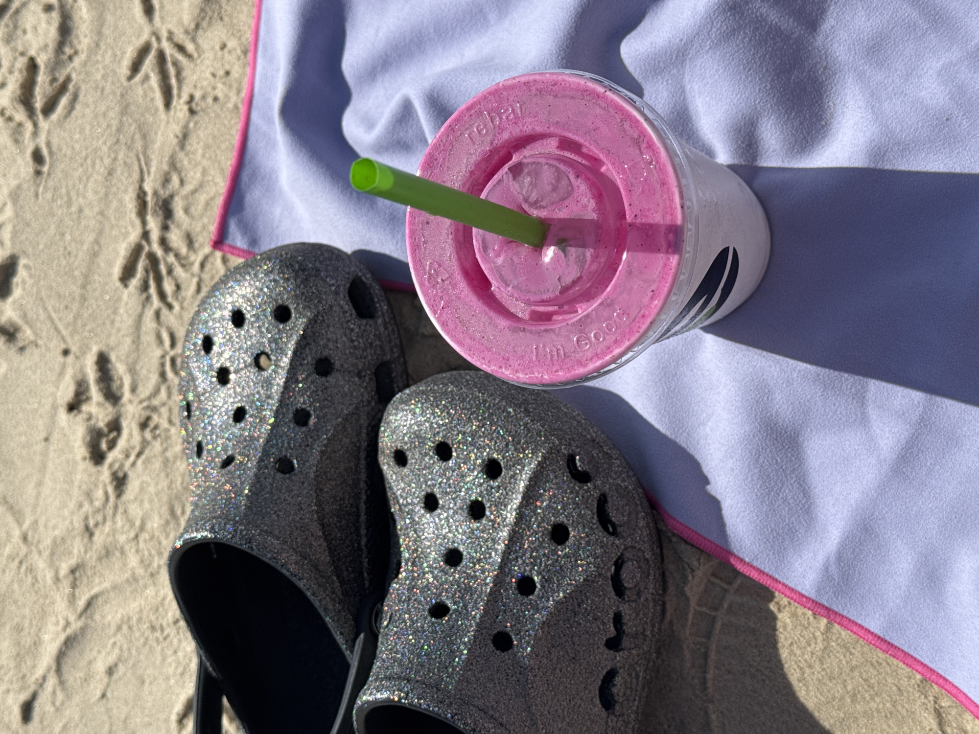 On a sunny beach in Tel-Aviv, Israel, a pair of silver glitter Crocs sit beside a light purple towel with a pink border. Next to the Crocs on the towel, is a white plastic cup with a clear lid containing a pink drink and a green straw. Bird footprints are visible in the sand.