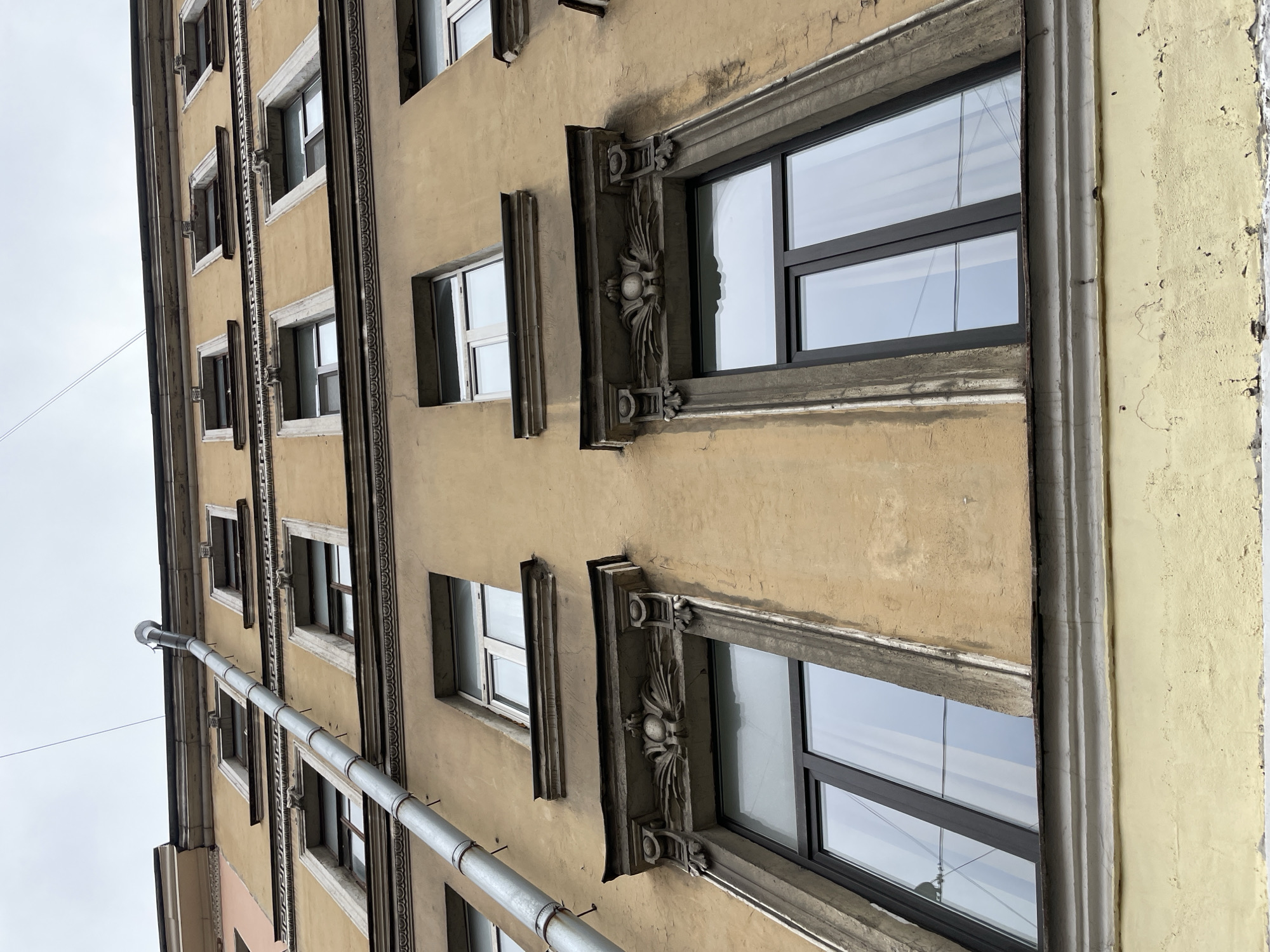 The image shows a multi-story building in Saint Petersburg, Russia. The building has a weathered, yellow-beige facade with ornate window frames. The windows are modern, with a double-paned glass design and black frames. A metal downspout runs vertically along the building's edge. The sky is visible in the reflection of the windows.