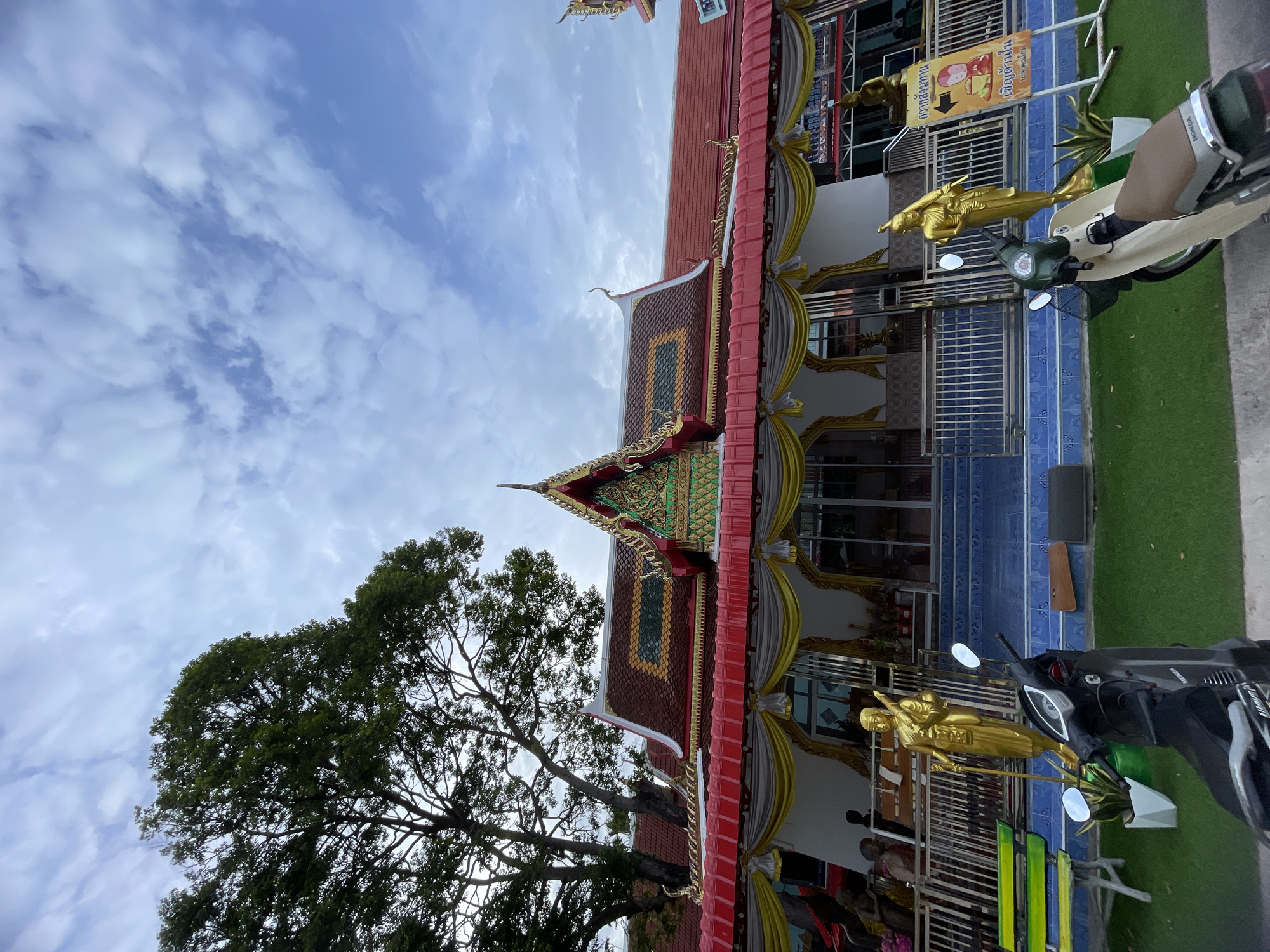 In Pattaya City, Thailand, a Buddhist temple is depicted under a cloudy sky. The temple features a prominent red tiled roof with intricate gold and green ornamentation on its gables. Red awnings with gold and white drapes extend from the building. Two golden statues of Buddhist figures stand outside the temple entrance, one on each side of a metal fence. In the foreground, two scooters are parked on a patch of artificial grass. To the right, a yellow sign with Thai script and an image of a monk is visible. A large tree with dense green foliage is situated to the left of the temple. Several individuals are visible inside and around the temple, some seated on a colorful bench.