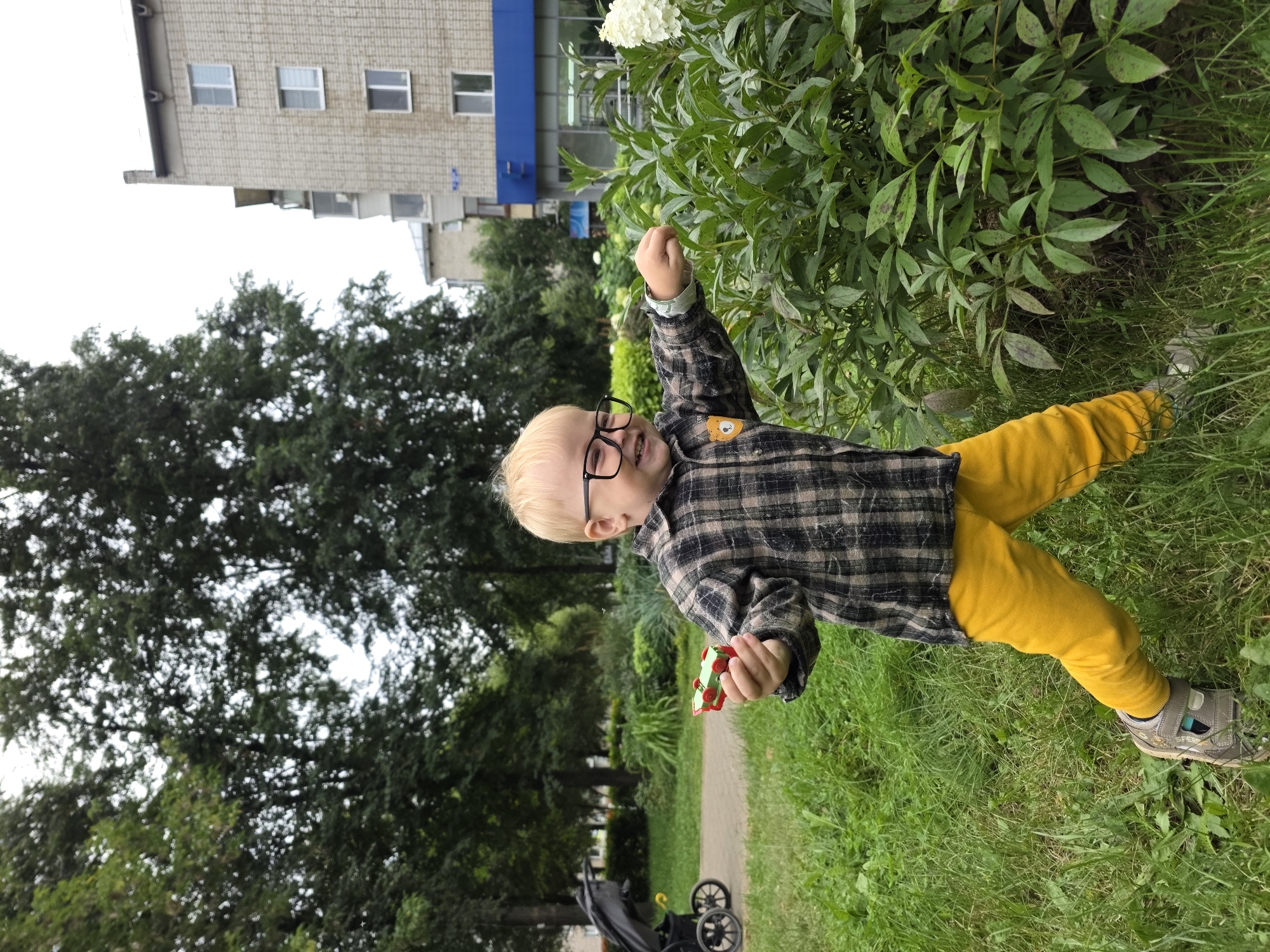 A young boy with blonde hair and glasses stands in a park, holding a small toy train. He is wearing a plaid shirt and yellow pants. He is reaching out towards a bush with white flowers. In the background, there is a stroller, trees, and a multi-story building. The scene appears to be in a residential area of Chaikovsky, Russia, possibly near a playground or courtyard.