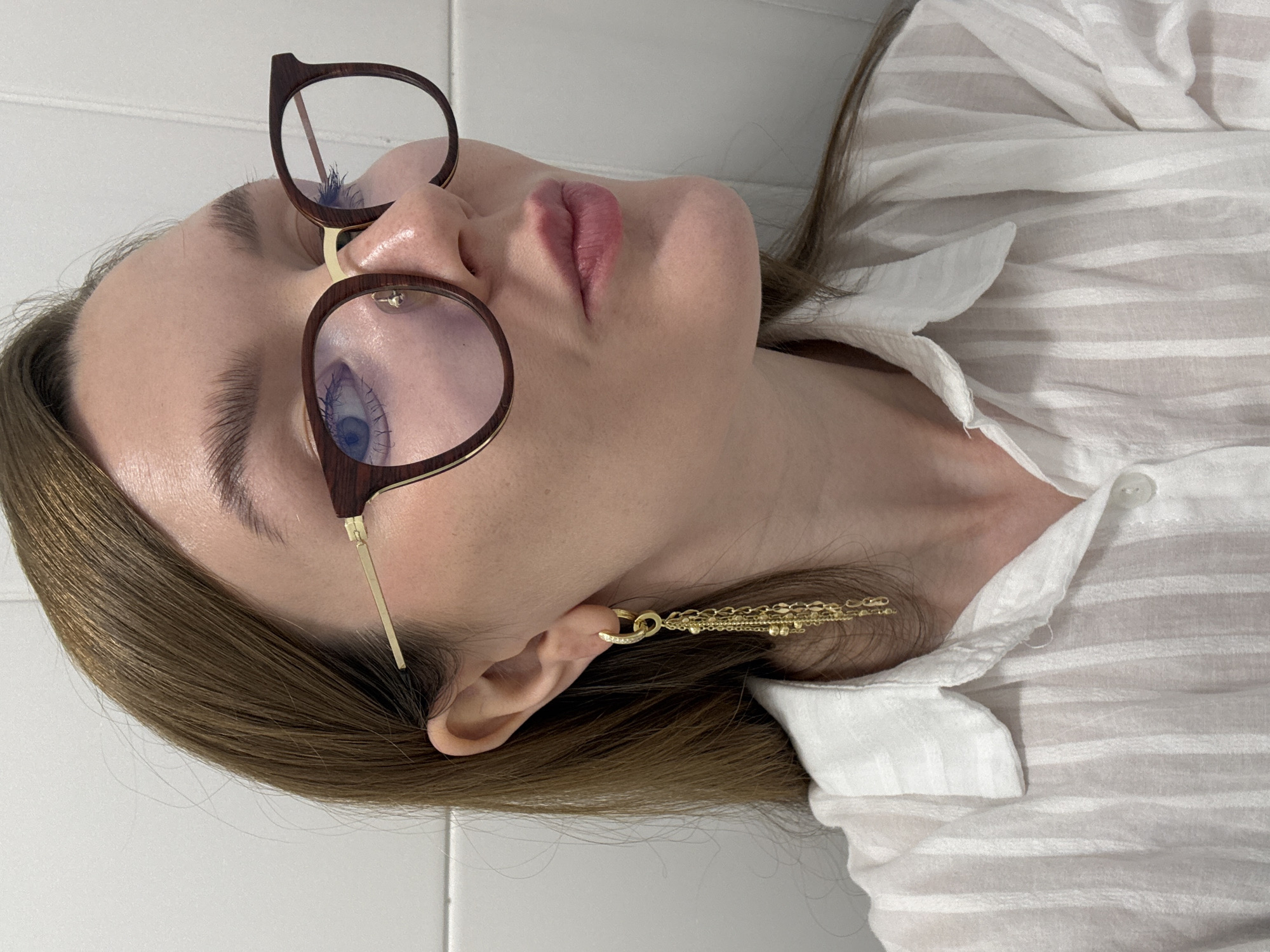 A medium close-up shot shows a woman from the chest up, facing right, against a white tiled wall. The woman has brown hair, is wearing large, brown-rimmed glasses with gold accents, and a white, vertically striped button-down shirt. She has a long, gold earring with multiple chains hanging from her right ear. Her lips are slightly parted and colored with a pink lipstick. The background consists of a white tiled wall with vertical grout lines. There is no visible indication of the location within the city of Izhevsk, Russia.
