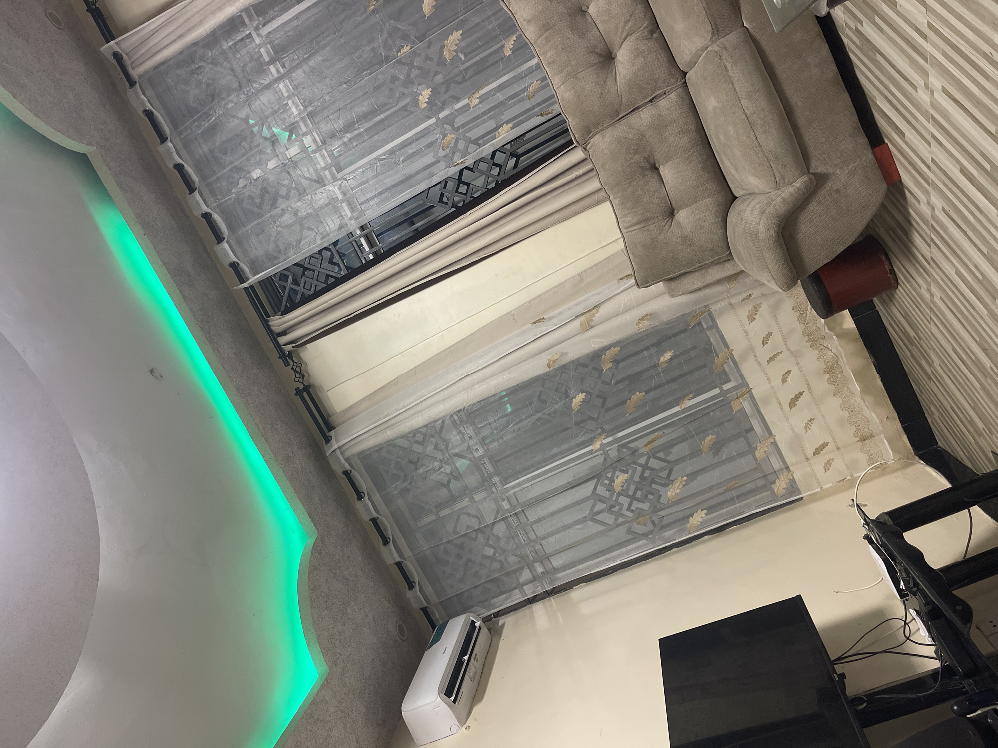 The image depicts a section of a living space, showing part of a couch, a window with sheer curtains, a wall with a green LED light, and an air conditioning unit. There are no people visible.
Object details: The couch is light brown, with tufted cushions. The sheer curtains have a repeating leaf pattern. The air conditioning unit is white and mounted near the floor. A black television stand and TV are in the bottom center. The wall adjacent to the couch has striped wallpaper.
Environment: The light suggests an indoor setting, likely during daylight hours.
No specific location details confirm Dar es Salaam, Tanzania.