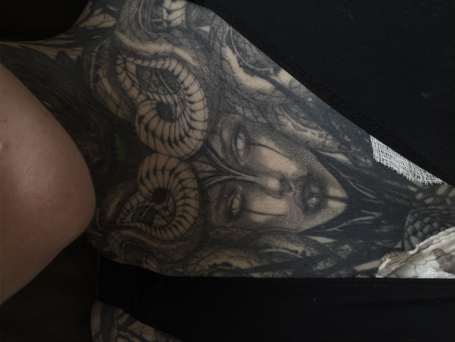 The image depicts a close-up view of a person's heavily tattooed neck and upper chest. The visible skin on the chin and jawline is fair-toned, contrasting with the extensive black and grey tattoo. The primary subject of the tattoo is a stylized female face with prominent, empty white eyes, dark tear-like streaks below the eyes, and a patterned design suggestive of snakes or ram horns forming a headdress. Intricate dark patterns surround the central face, extending across the throat and onto the upper chest and shoulders. A small area of green coloration is visible within the tattoo design near the upper center. The individual is wearing a dark, possibly black, garment with a smooth texture that frames the tattooed area on both sides. A portion of a lighter, patterned fabric, resembling camouflage, is visible on the lower left, with a small section of white mesh-like material further to the right. The background is dark and out of focus. The specific location in the city of Chaikovsky, Russia, cannot be determined from the visual information provided in the image.