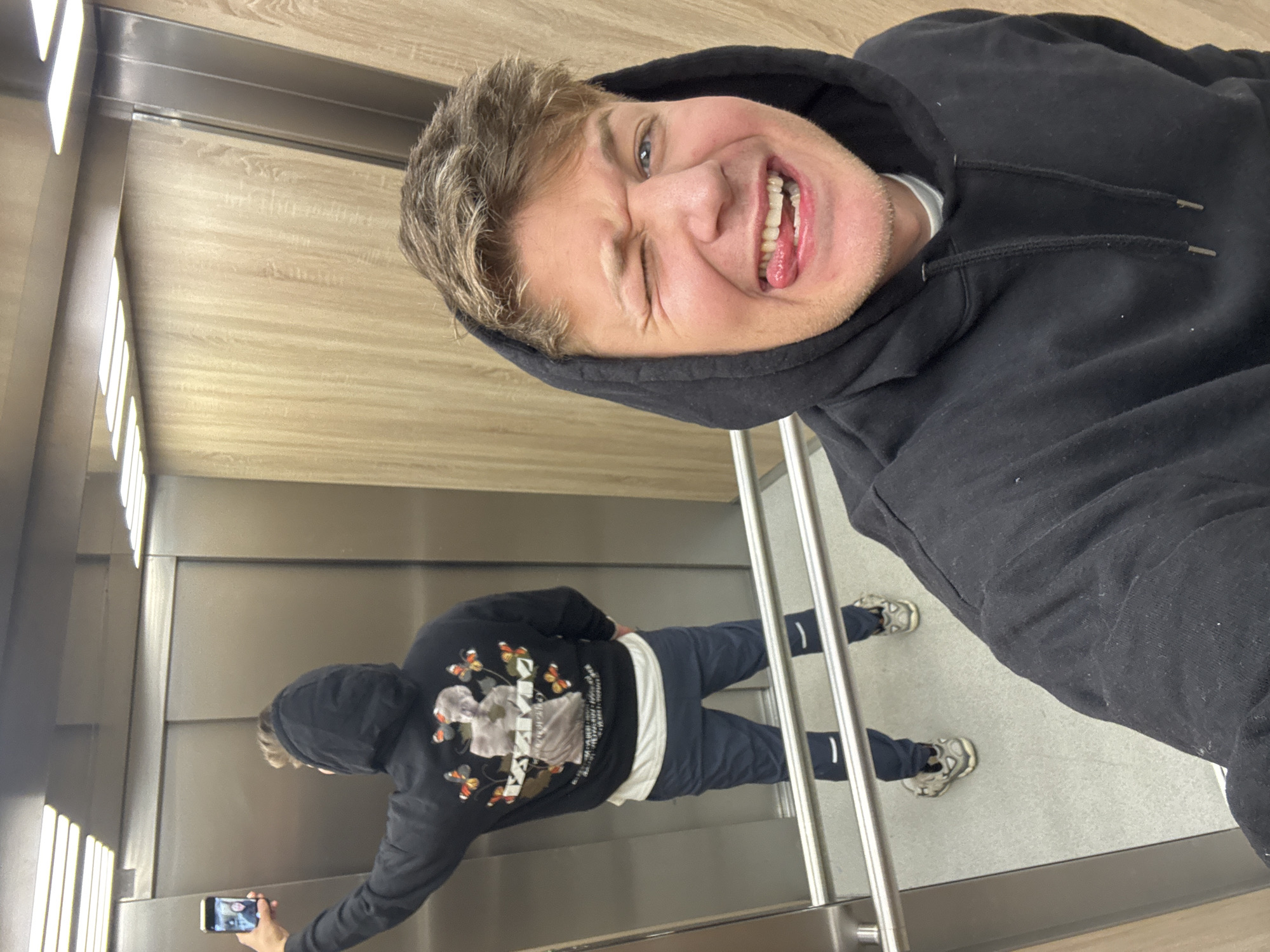 The image captures a playful mirror selfie taken inside an elevator. The main subject is a young man, rotated 90 degrees counter-clockwise in the frame, appearing to lie horizontally. He has light brown, slightly wavy hair and blue eyes, and is wearing a black hooded sweatshirt with the hood up. He is making a humorous face, winking his left eye, sticking out his tongue, and displaying a wide, cheerful grin that shows his teeth. A faint stubble is visible on his face.
The elevator interior features wood-paneled walls with a light brown, vertical grain on one side, and metallic, possibly stainless steel, surfaces on the opposite side where a large mirror reflects the man's full body. The floor is a plain gray. Vertical strip lights are visible on the metallic wall, reflecting in the mirror.
In the mirror's reflection, the man is seen from behind and slightly to the side, standing upright. He holds a smartphone in his right hand, pointed towards the mirror to capture the selfie. His black hoodie features a prominent white graphic on the back depicting what appears to be a classical statue, possibly a bust, surrounded by multiple colorful butterflies. Below this graphic, there are several lines of faint, unreadable white text. He is also wearing dark blue trousers and white and beige athletic shoes.
The overall mood is lighthearted and cheerful. The scene is set in an elevator, specifically located in Plovdiv, Bulgaria.