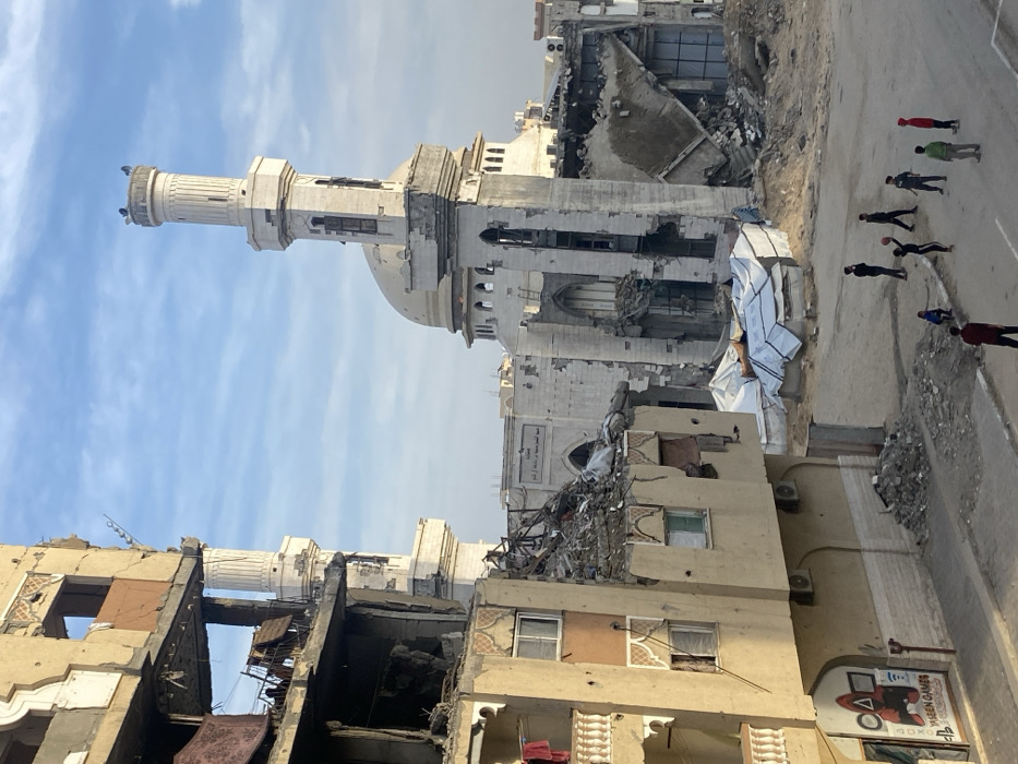 The image displays a street scene in Khan Yunis, Palestinian Territory, characterized by extensive structural damage under a partly cloudy sky. On the left, a multi-story building shows significant partial collapse on its upper levels, with exposed concrete and internal fixtures. A lower balcony on this structure has laundry hanging. The ground floor features a storefront labeled "Yaseen Games" with a "Squid Game" themed graphic. Dominating the midground is a large mosque complex, heavily damaged. Its prominent minaret exhibits structural compromise and visible openings, while the main mosque structure behind it has large sections collapsed, revealing internal spaces and debris. A central dome on the mosque appears partially intact but scarred. Piles of rubble are present throughout the area, with some covered by white tarpaulins near the mosque. The street surface is unpaved and scattered with debris. In the foreground, approximately eight to nine individuals, primarily children and young males, are gathered on the street, with one actively dribbling a basketball, suggesting a game in progress.