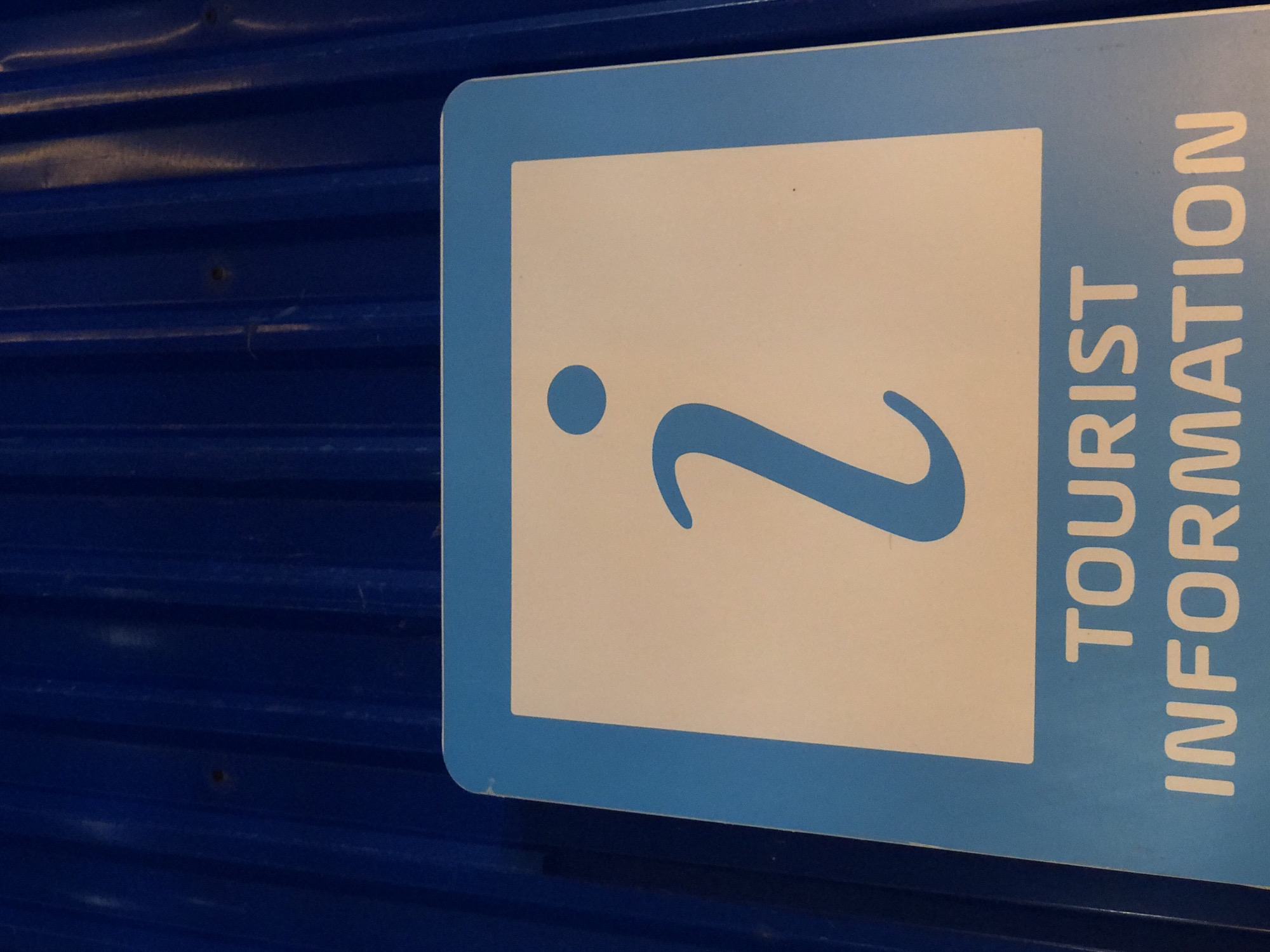 The image shows a close-up of a blue and white sign with the international "i" symbol and the words "TOURIST INFORMATION" in white, sans-serif font. The sign is mounted on a ribbed, dark blue surface. The lighting suggests it might be outdoors or in a well-lit indoor area. The sign is oriented vertically. The context of Đà Nẵng, Vietnam, is not explicitly visible in the image itself; the description is solely based on the visual elements present.
