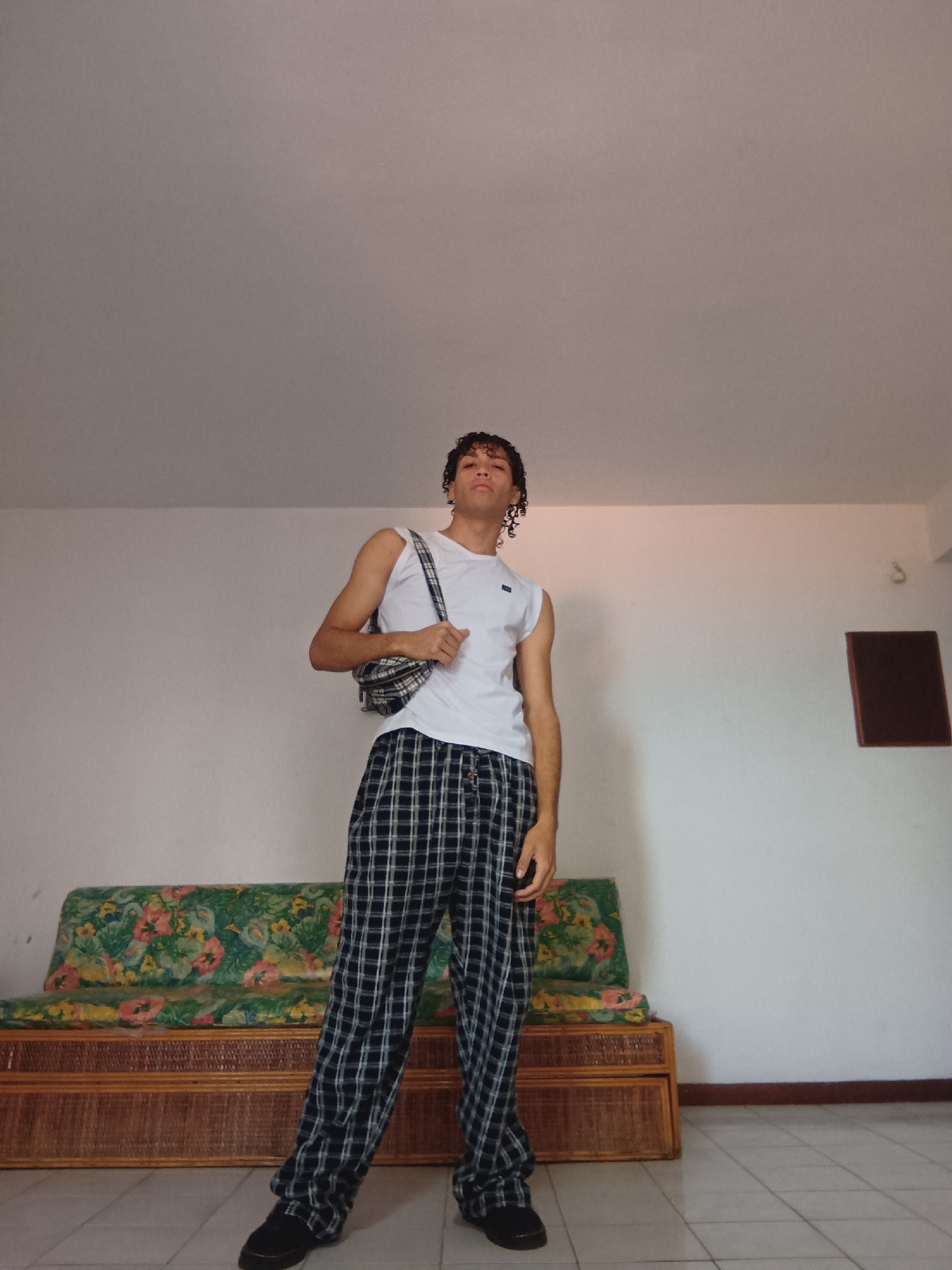 The image displays a young person, possibly male, with dark curly hair, standing indoors and posing for the camera. They are wearing a white sleeveless top, black and white plaid wide-leg pants, and dark-colored shoes. A plaid shoulder bag, matching the pattern of the pants, is slung across their body. The person has a neutral to slightly confident expression and is looking directly at the viewer.
The setting is an indoor room with plain white walls and a light-colored tiled floor. To the left and behind the subject, there is a long wooden sofa with a woven or rattan base, topped with a vibrant floral cushion featuring green, orange, pink, and yellow patterns. On the wall to the right of the subject, a small, dark brown rectangular object is mounted. The lighting suggests it is daytime. The scene appears to be a casual indoor environment, likely a living area within a residence in Porlamar, Venezuela. No visible text is present in the image.