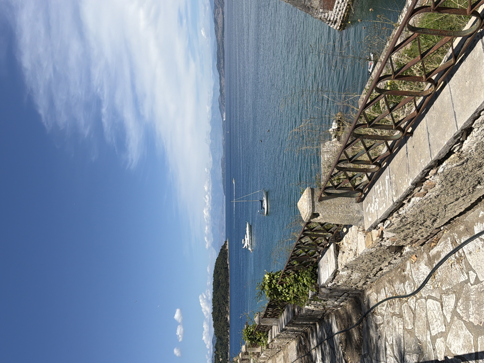 This media file captures a serene coastal landscape in Corfu, Greece, under a clear, bright sky. The dominant feature is a vast expanse of calm, blue-green sea. In the distance, a long, dark, undulating landmass, appearing hazy and possibly mountainous, stretches across the horizon.
The sky, particularly on the left, is adorned with scattered white, wispy clouds, fading to a clearer blue towards the right. On the tranquil water, two white boats are visible in the mid-ground: one sailboat with a prominent mast and a smaller, sleek powerboat or yacht. A very small, dark boat is also discernible closer to the right-hand shore.
In the foreground, a weathered stone and concrete wall runs diagonally, partially enclosing the scene. This wall shows signs of age, with some crumbling sections and visible block work. Mounted atop it is a rusty, dark brown metal railing, featuring geometric patterns and distinct oval sections. Patches of green vegetation, including bushes and dry grass, grow on and around this structure. A black electrical cable is visible running along the lower part of the wall. Faint, illegible graffiti or markings can be seen on the concrete surface of the wall.
The time of day appears to be late morning or early afternoon, indicated by the bright, overhead sunlight. The weather is clear and sunny, contributing to the overall peaceful and picturesque atmosphere. No people are visible, enhancing the sense of quietude in this beautiful Mediterranean setting.