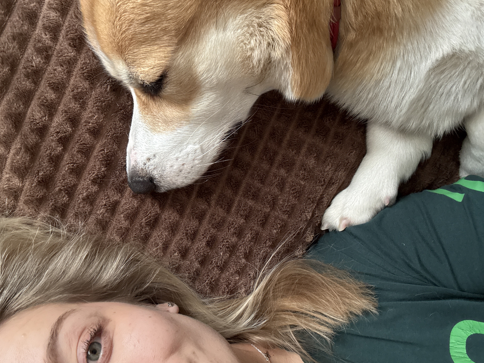 A close-up indoor shot shows a woman with blonde hair lying on a brown textured surface. A corgi dog, with a tan head and white body, is resting its head and paw on the same surface. The woman is wearing a dark green shirt with light green lettering. A small piece of jewelry is visible around her neck. The specific location of this image is not determined.