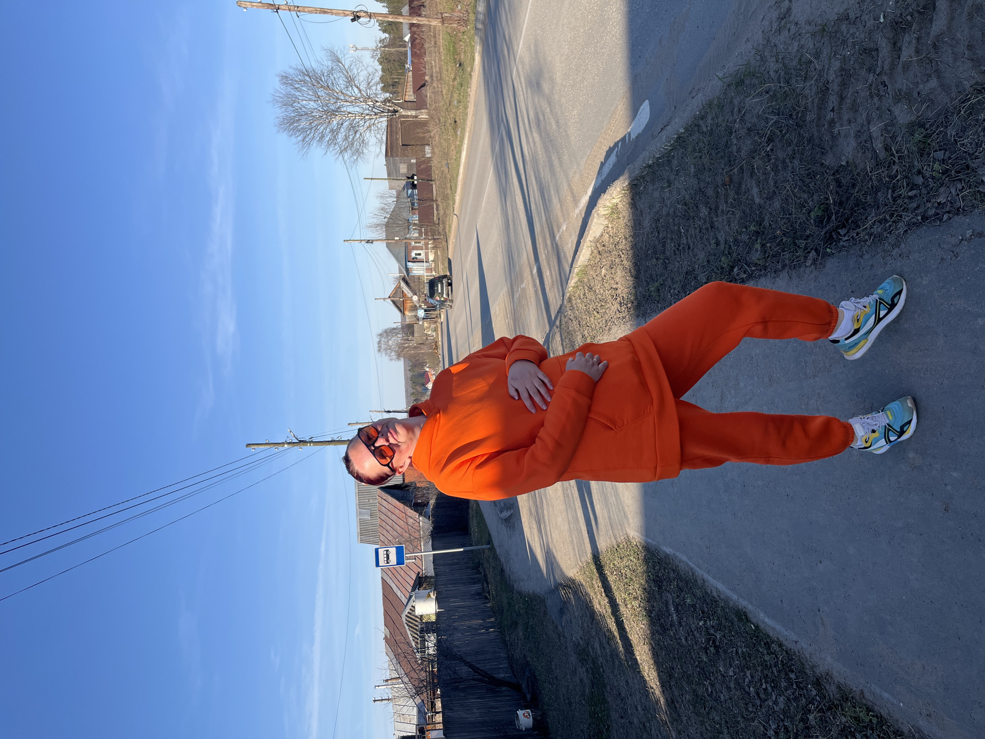 The image captures a person standing on a sunny day in a semi-rural or village setting in Chaikovsky, Russia. The individual, appearing to be an adult, is dressed in a bright orange tracksuit and dark sunglasses, with colorful sneakers that are primarily blue, yellow, and white. They are standing on a paved surface, possibly a sidewalk or road shoulder, with their right hand placed on their stomach.
Behind the person, a paved road stretches into the distance, flanked by grassy verges and wooden fences. Scattered houses and buildings, some with a rustic appearance, line the road, and bare trees suggest early spring or late winter. In the background, what appears to be more open land or forest is visible. Overhead, electrical poles with wires are prominent against a clear, vibrant blue sky, indicating bright daylight. The sun casts distinct shadows on the ground to the left of the person.
A notable detail is a blue and white bus stop sign visible behind and to the left of the person, featuring a universal white bus icon. The overall atmosphere is bright and calm, capturing a casual outdoor moment.