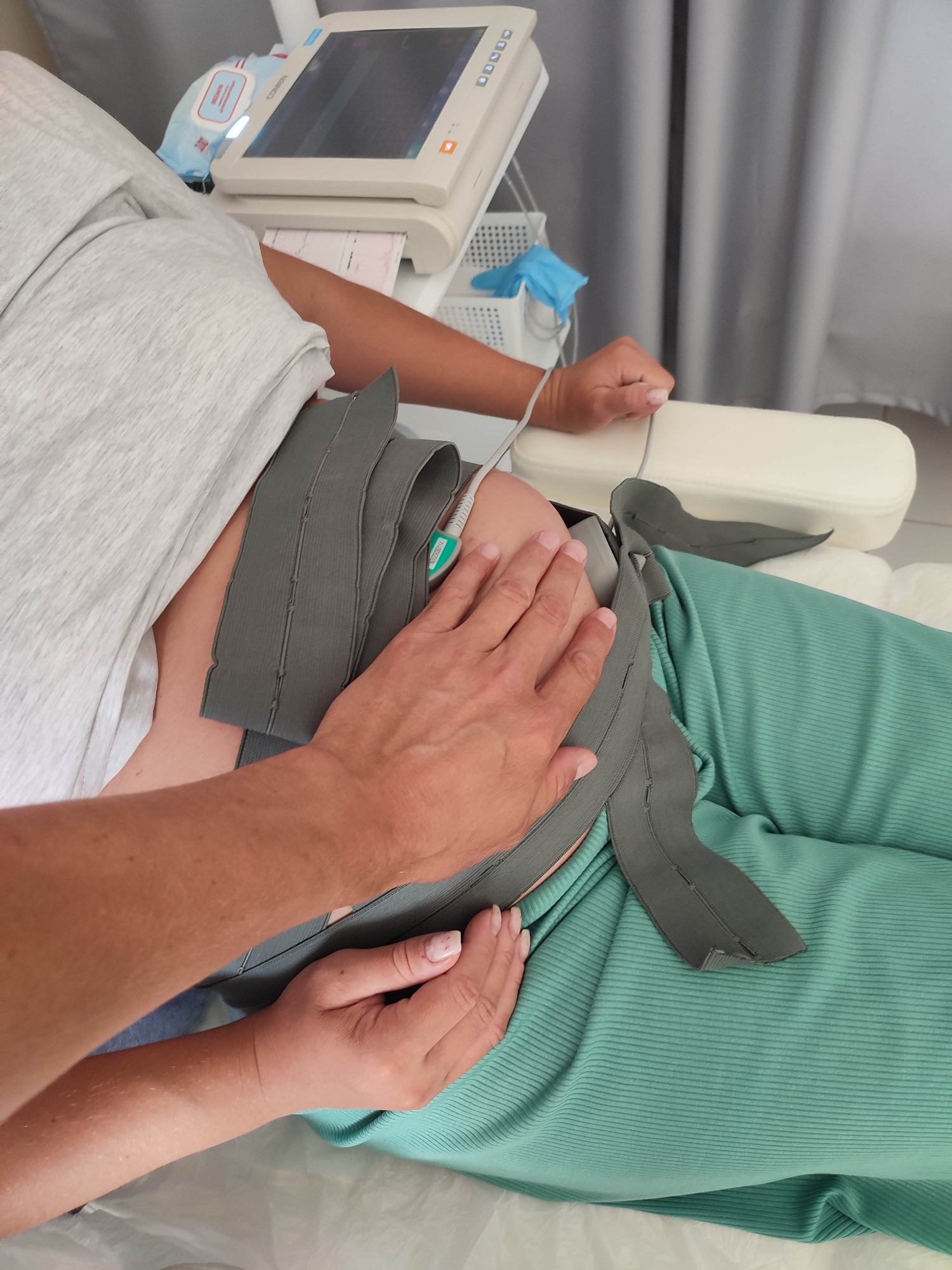In an indoor medical setting, a person is lying down, wearing a light grey t-shirt and teal ribbed pants. A grey medical strap is around their abdomen, connected to a monitor visible in the background. Another person's hands are gently resting on the strap and the abdomen, suggesting a caring or monitoring interaction. The background includes a medical monitor displaying a strip chart, a white plastic basket with medical supplies like wipes and gloves, and a grey curtain. The lighting is neutral, and the perspective is a close-up shot focusing on the abdominal area and the hands.