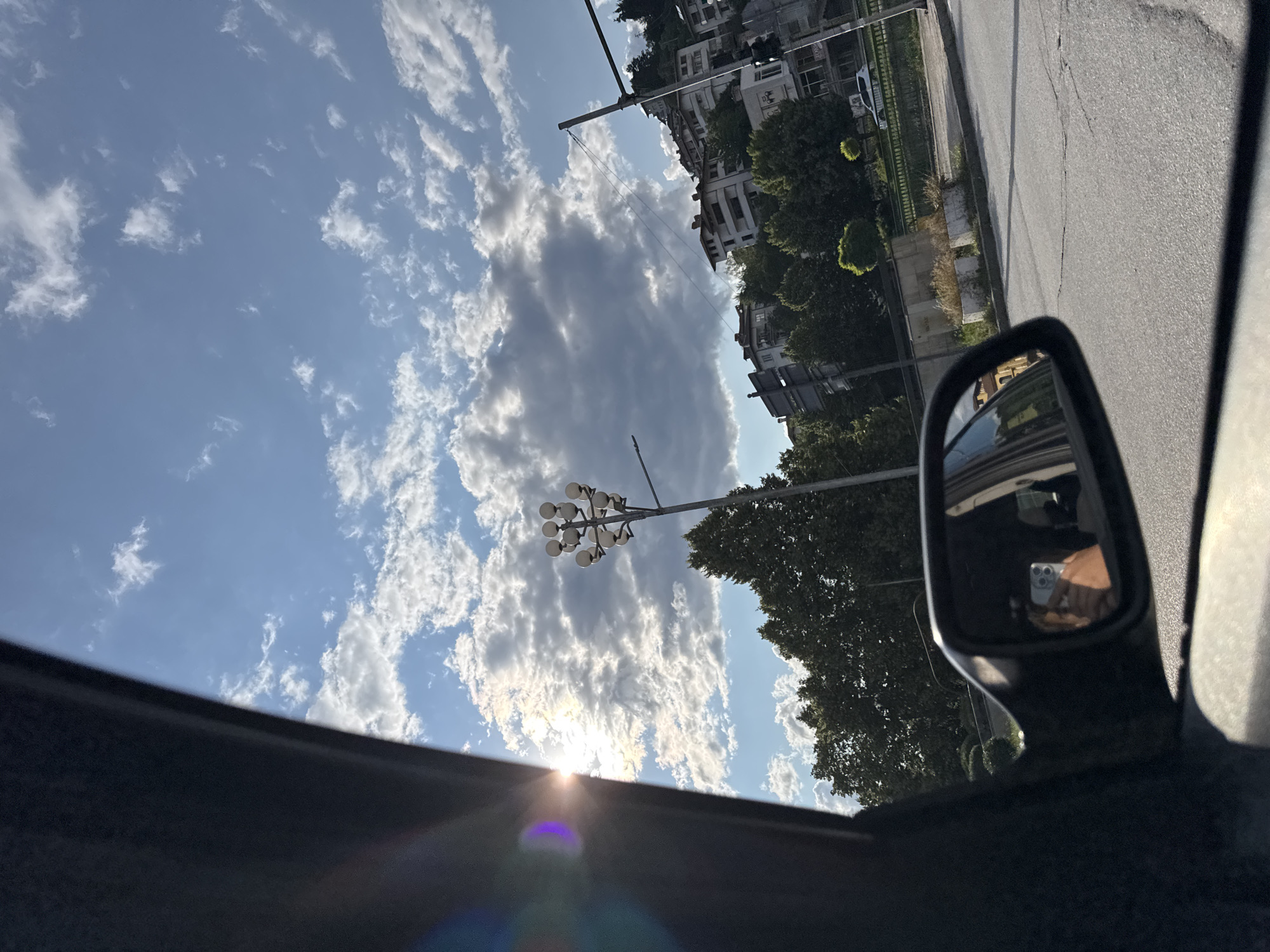 The image is taken from inside a vehicle, looking upwards and to the right, with the viewpoint rotated approximately 90 degrees counter-clockwise. The primary subjects are the sky, buildings, trees, and a side-view mirror.
The sky is bright blue with scattered white and grey clouds. The sun is visible, low in the sky, casting a lens flare. The clouds are illuminated from behind by the sun, creating a dramatic contrast.
In the background, a street is visible with a paved surface. Along the street, there are several buildings with white facades and multiple stories, appearing to be residential or commercial structures. Lush green trees line the street between the buildings and the road, some with neatly trimmed bushes. A street lamp with multiple bulbs is also visible.
A side-view mirror of the vehicle is prominently featured on the right side of the frame. Reflected in the mirror is the interior of the vehicle and a person's hand holding a smartphone, which is angled to capture a selfie.
The overall scene suggests it is daytime, likely late afternoon or early evening due to the low sun angle. The weather appears to be clear with some cloud cover. The location context is Smolyan, Bulgaria, indicated by a sign that is not clearly legible in the main image but visible in cropped images. The mood is casual, perhaps a drive through a town on a pleasant day.
