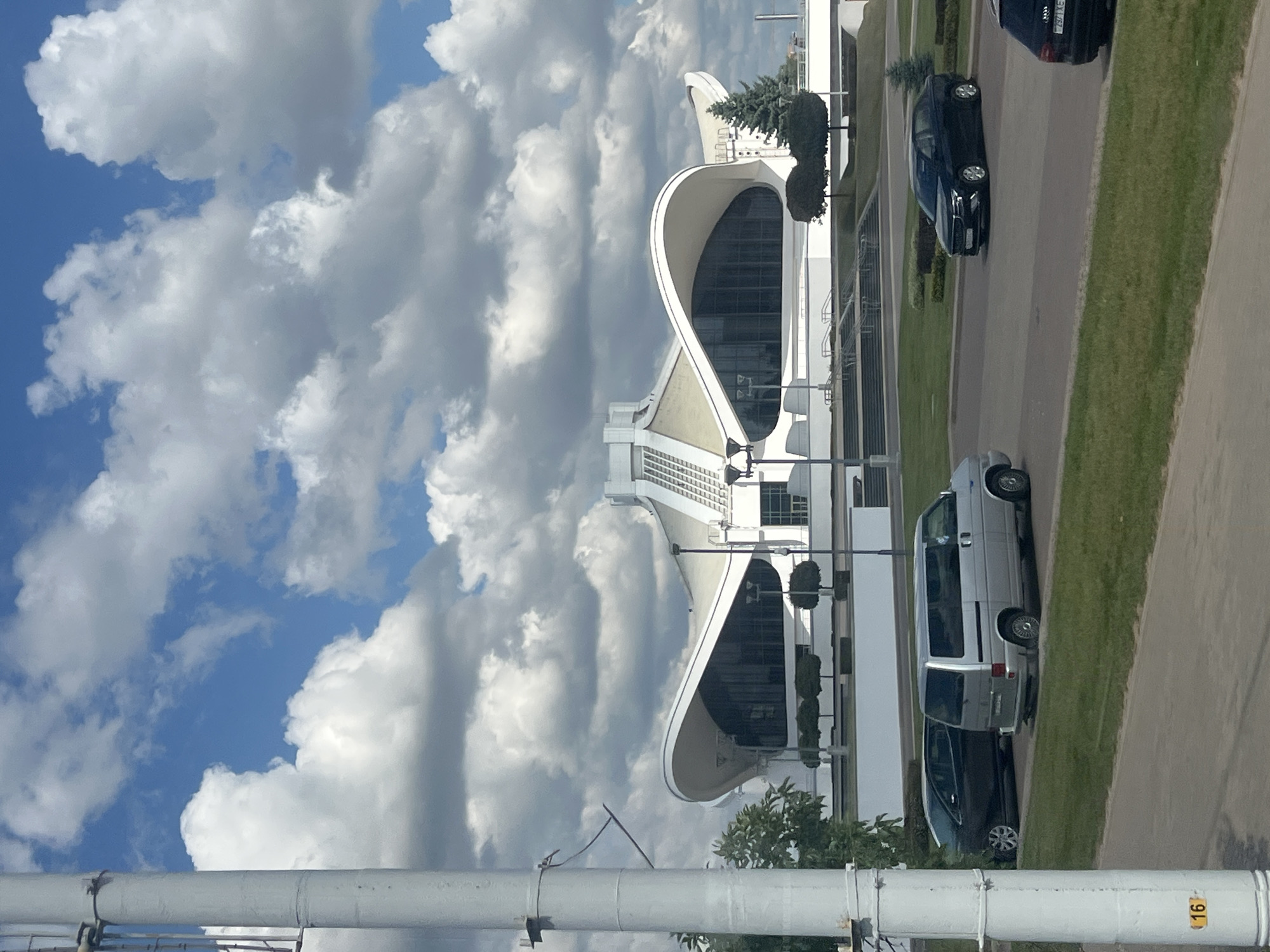 The image captures the Palace of Sports in Minsk, Belarus, on a partly cloudy day. In the foreground, a light-colored pole with a yellow marker "16" is visible on the left side. A silver van and a dark sedan are parked on a paved area with a grassy median. Behind them, the distinct white, wing-like structure of the Palace of Sports dominates the background. The building features large, dark-tinted windows and a modern architectural design. Several trees, some pruned into spherical shapes and a few evergreen trees, are strategically placed around the building and the surrounding landscape. The sky is a vibrant blue with scattered white, fluffy clouds. The scene appears to be a street view from a vehicle, looking towards the sports venue.