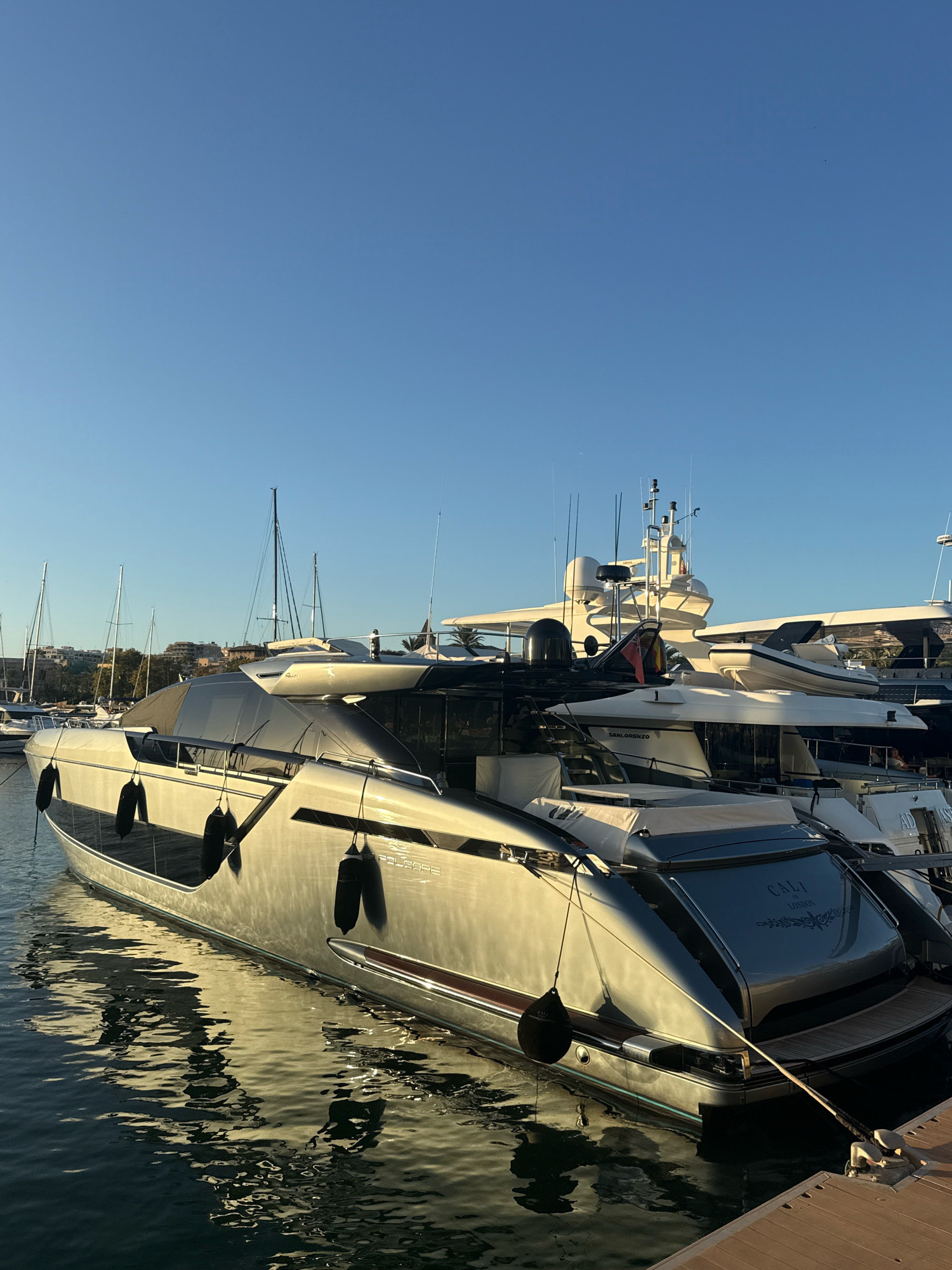 The image captures a sleek, metallic-grey yacht named "CALI OF LONDON" docked in a harbor in Palma, Spain. The yacht is the primary subject, positioned in the foreground and angled slightly towards the right of the frame. Its polished surface reflects the clear blue sky and the gentle ripples of the water. Several black fenders hang along its hull, indicating it is secured to the dock.
In the background, other boats, including sailboats with tall masts, are visible, suggesting a busy marina. The architecture of buildings and palm trees can be seen in the distance, hinting at the urban landscape of Palma. The lighting suggests it is either early morning or late afternoon, with the sun casting a warm glow on the yacht and its surroundings. There are no people visible in the image. The overall scene depicts a serene and luxurious maritime environment.