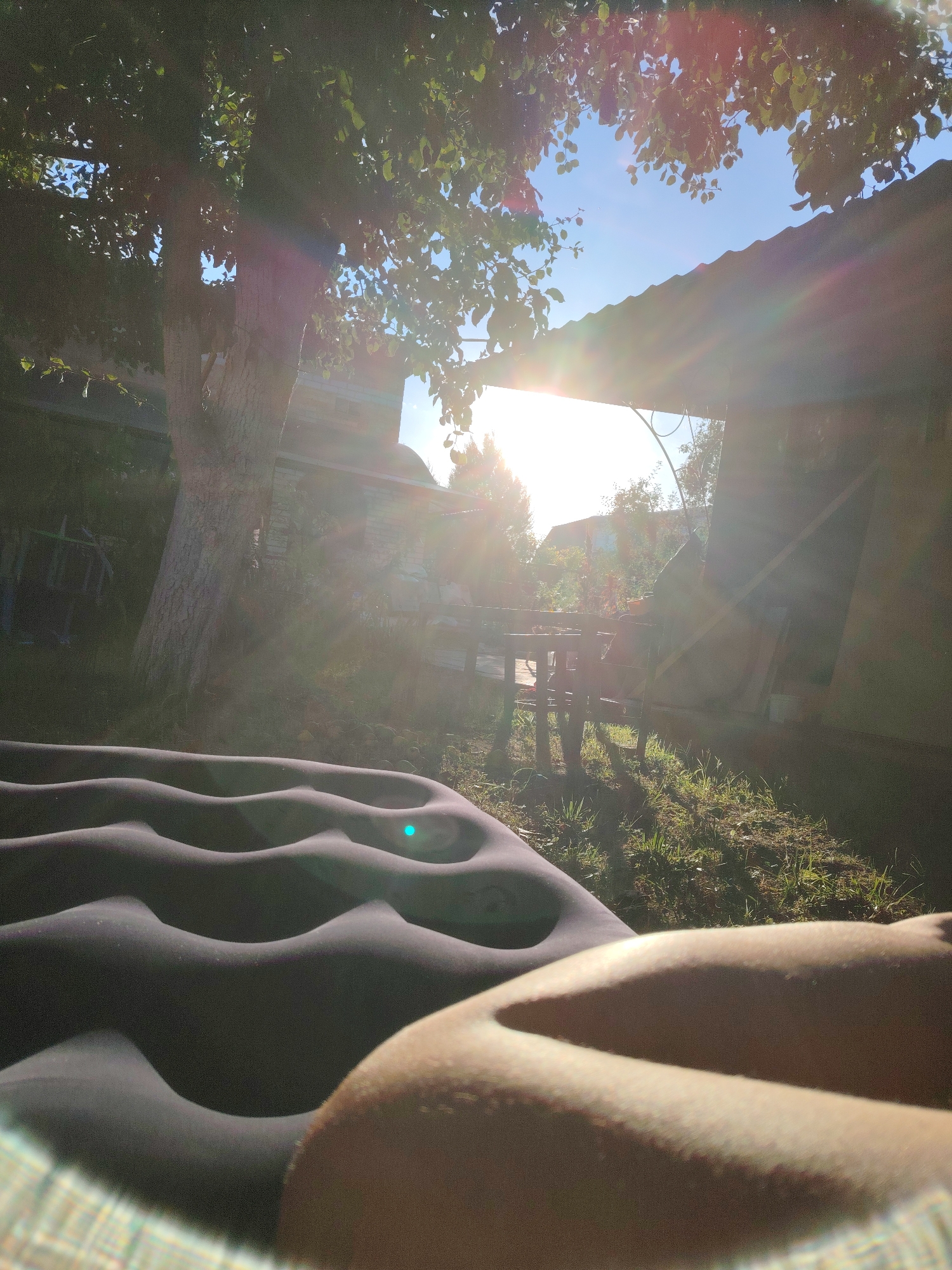 In an outdoor setting, bathed in bright sunlight and lens flare, an inflated air mattress dominates the foreground. The mattress is dark gray and has a distinct wavy pattern. To the right, a person's bare legs and knee are visible, resting on the mattress. The background depicts a yard with a large tree on the left, its branches casting shadows. Sunlight streams through the leaves and the opening of a roofed structure. A wooden table and chairs are visible behind the air mattress, and further in the background, buildings and trees can be seen under a clear blue sky. The overall impression is a relaxed, daytime scene.