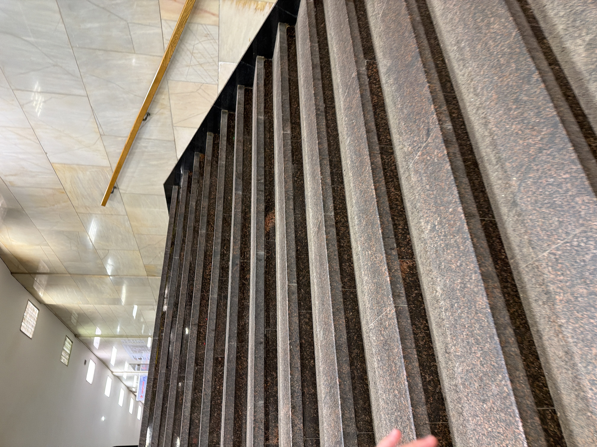 The image shows a view looking down a stairway in the Moscow Metro. The steps are constructed from a reddish-brown granite material. The walls are clad in light-colored marble. A wooden handrail is visible on the left. The perspective indicates the photo was taken from an elevated position, looking down the stairwell toward the lower level. Fluorescent lights are visible on the ceiling of the lower level.