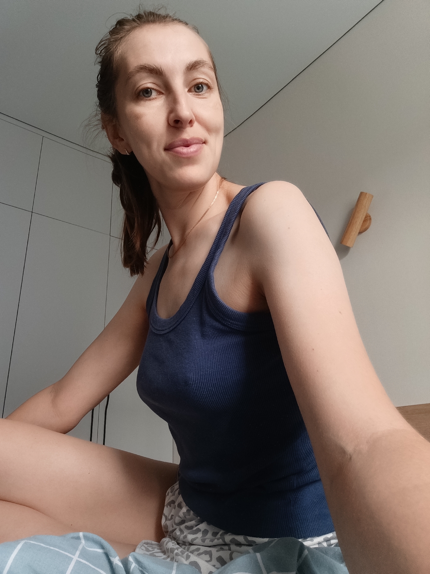 A woman with brown hair pulled back in a ponytail is sitting on a bed in a room. She is wearing a dark blue tank top and patterned shorts. Her legs are crossed. A duvet is visible at the bottom of the frame. The background shows a white closet and a grey wall with a wooden peg. The lighting is natural and diffused. There is no indication of the city of Chaikovsky, Russia in the image.