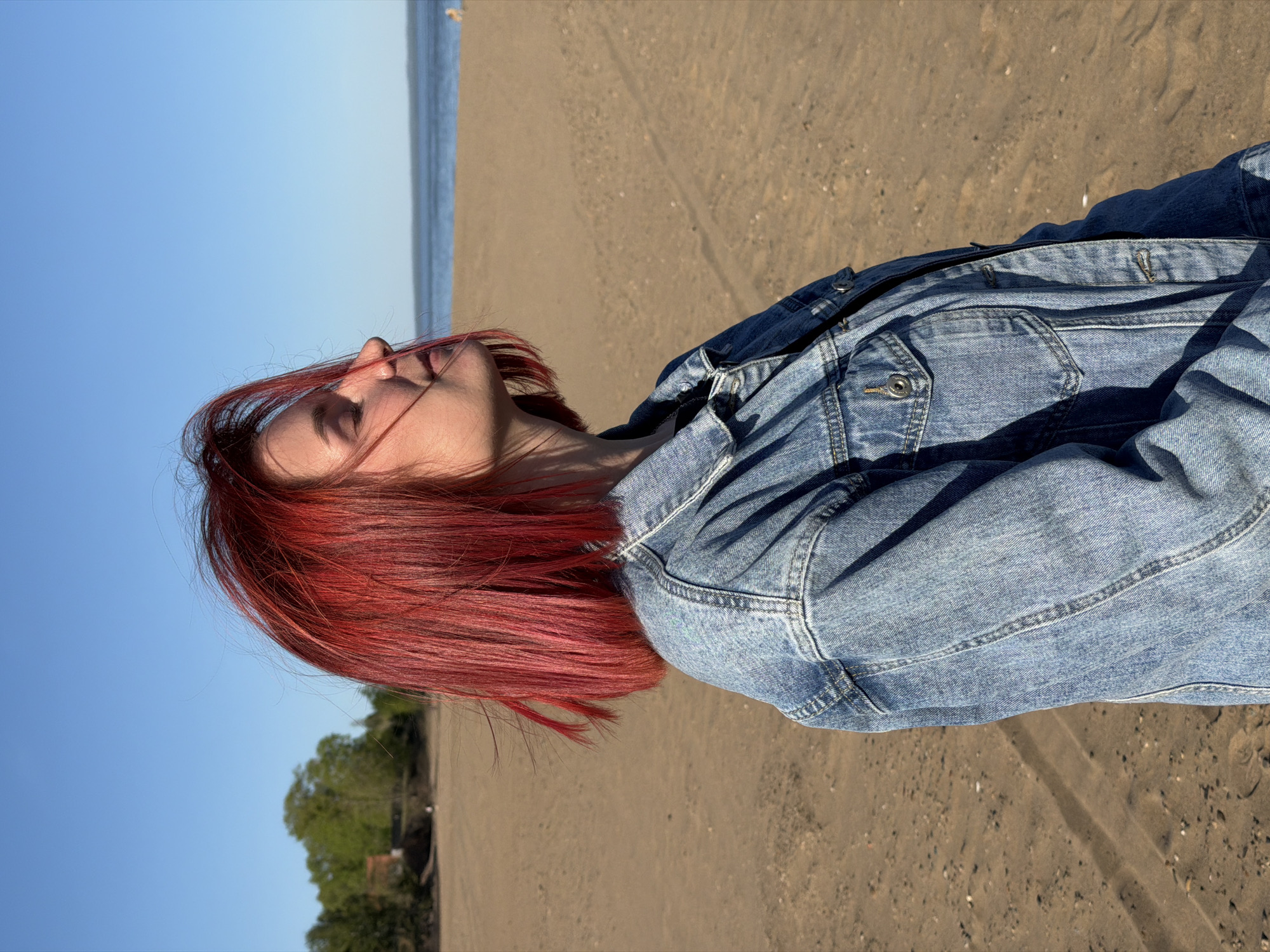 A young woman with vibrant red, short hair is lying on a sandy beach. She's wearing a denim jacket. The beach is likely located in Chaikovsky, Russia, given the prompt. A body of water, possibly a river or lake, is visible in the background along with patches of greenery. The sky is clear and blue, suggesting a sunny day. There are tire tracks in the sand. The woman's head is tilted back, possibly looking up at the sky.