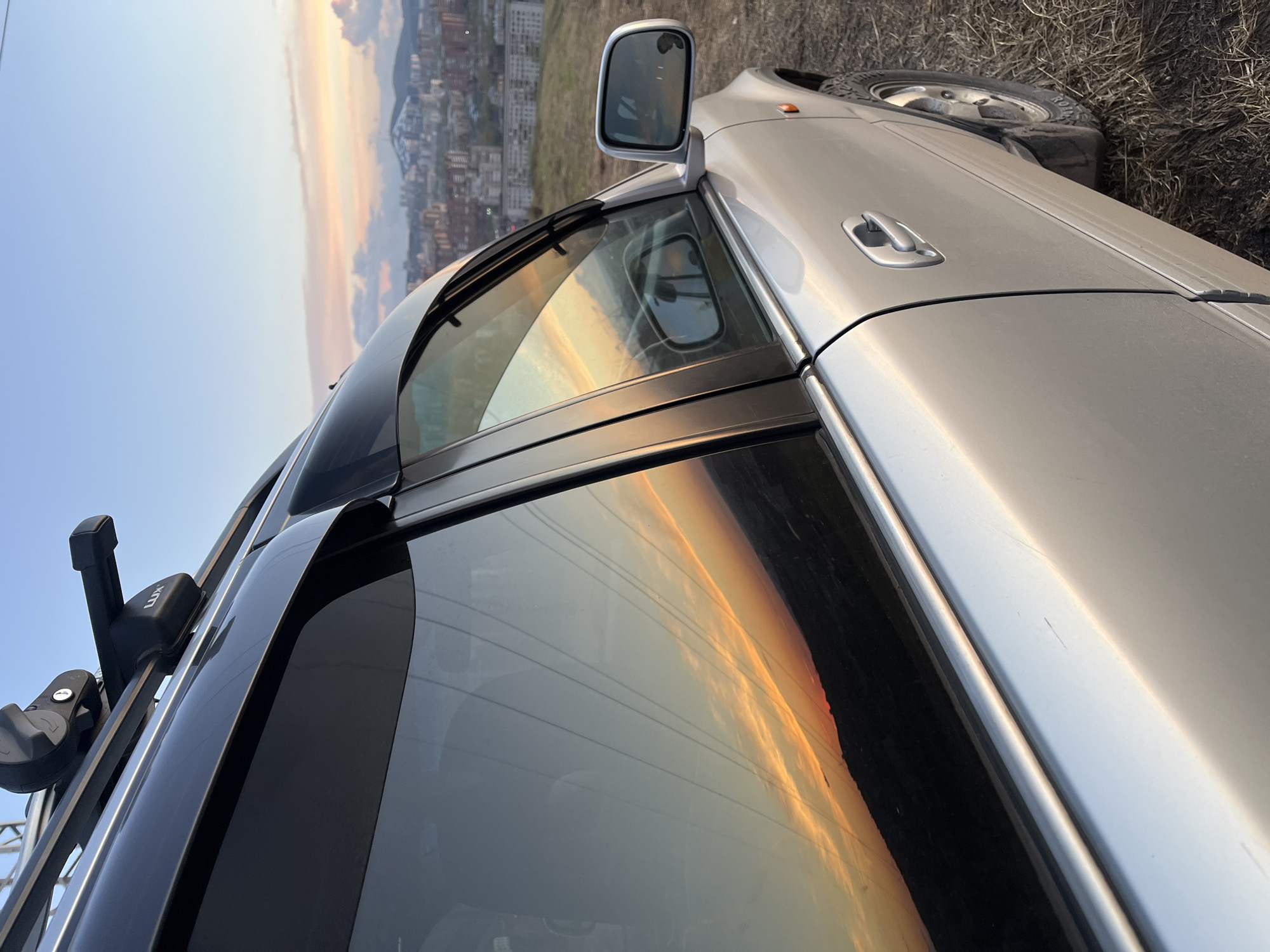 The image is a close-up shot of a silver car, possibly a van or SUV, parked near Krasnoyarsk, Russia. The car's side mirror, door handle, roof rack, and part of a tire are visible. The sunset and city skyline are reflected in the car's windows. The ground near the tire appears to be covered with dry vegetation.