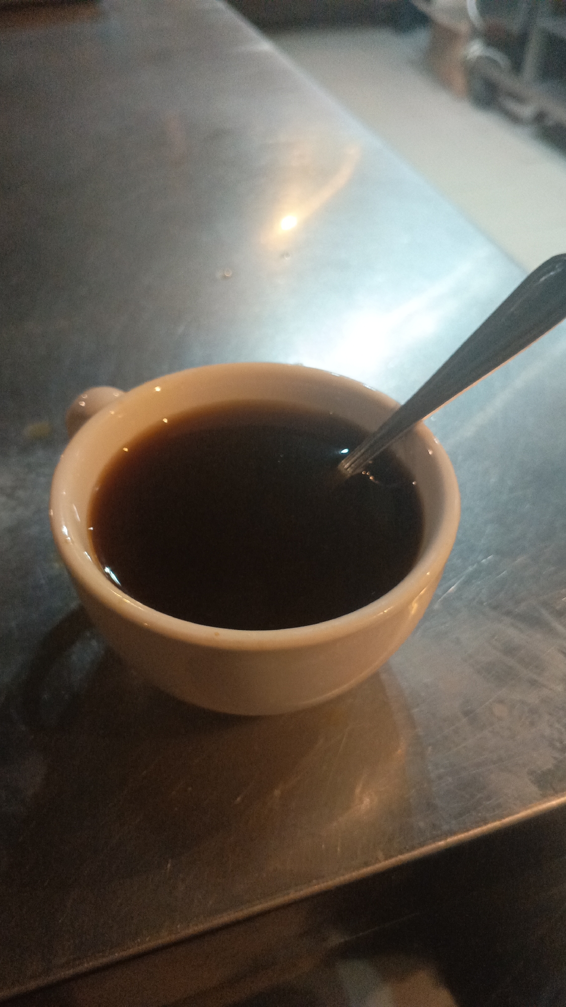 A close-up shot shows a white ceramic mug filled with dark liquid, likely coffee, resting on a metallic surface. A silver spoon is partially submerged in the liquid. The metallic surface reflects ambient light, creating highlights and shadows. The background is out of focus, hinting at an indoor environment with soft lighting. The scene is static, with no visible people or active events. The location is not discernible from the image itself and is stated to be in Mytishchi, Russia, without providing any visual evidence to support this.