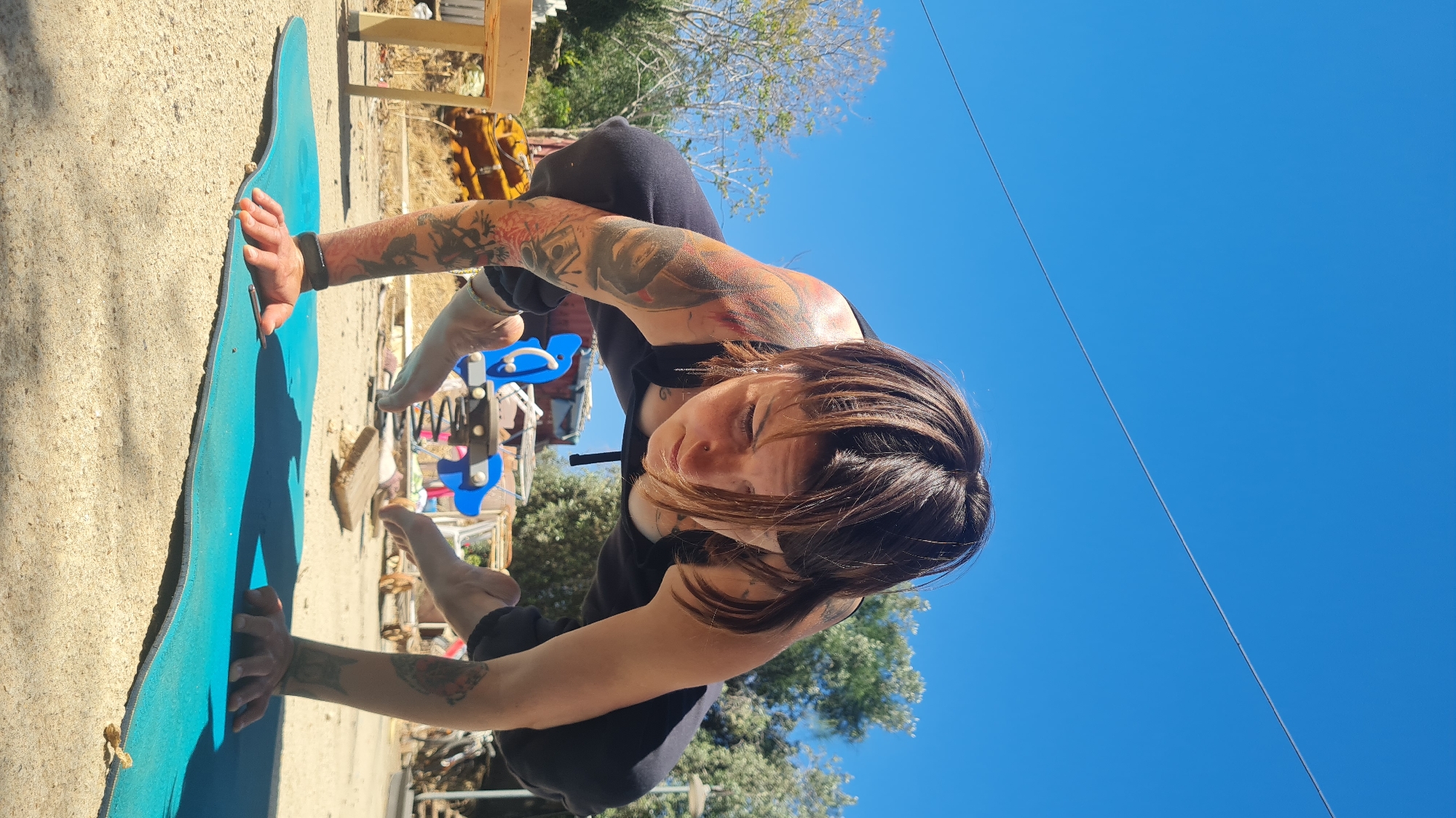 A woman with dark hair and visible tattoos on her arms is performing an arm balance pose, resembling a variation of Bakasana (Crane Pose), outdoors on a bright day. She is wearing dark athletic attire, including a sleeveless top and leggings. Her hands are placed firmly on a teal blue yoga mat, supporting her body as her legs are lifted and crossed behind her. Her expression is focused and calm.
The setting is an open-air area, likely in the Lachish Regional Council, Israel, under a clear, vibrant blue sky. The ground beneath the yoga mat is light-colored and appears to be sandy or gravelly. In the background, hints of trees and some rustic wooden structures are visible. To the right of the woman, a blue children's spring rider, shaped like an animal, can be seen, suggesting a park or backyard environment. The strong sunlight indicates a daytime scene with clear weather, casting defined shadows. There is no visible text in the image.