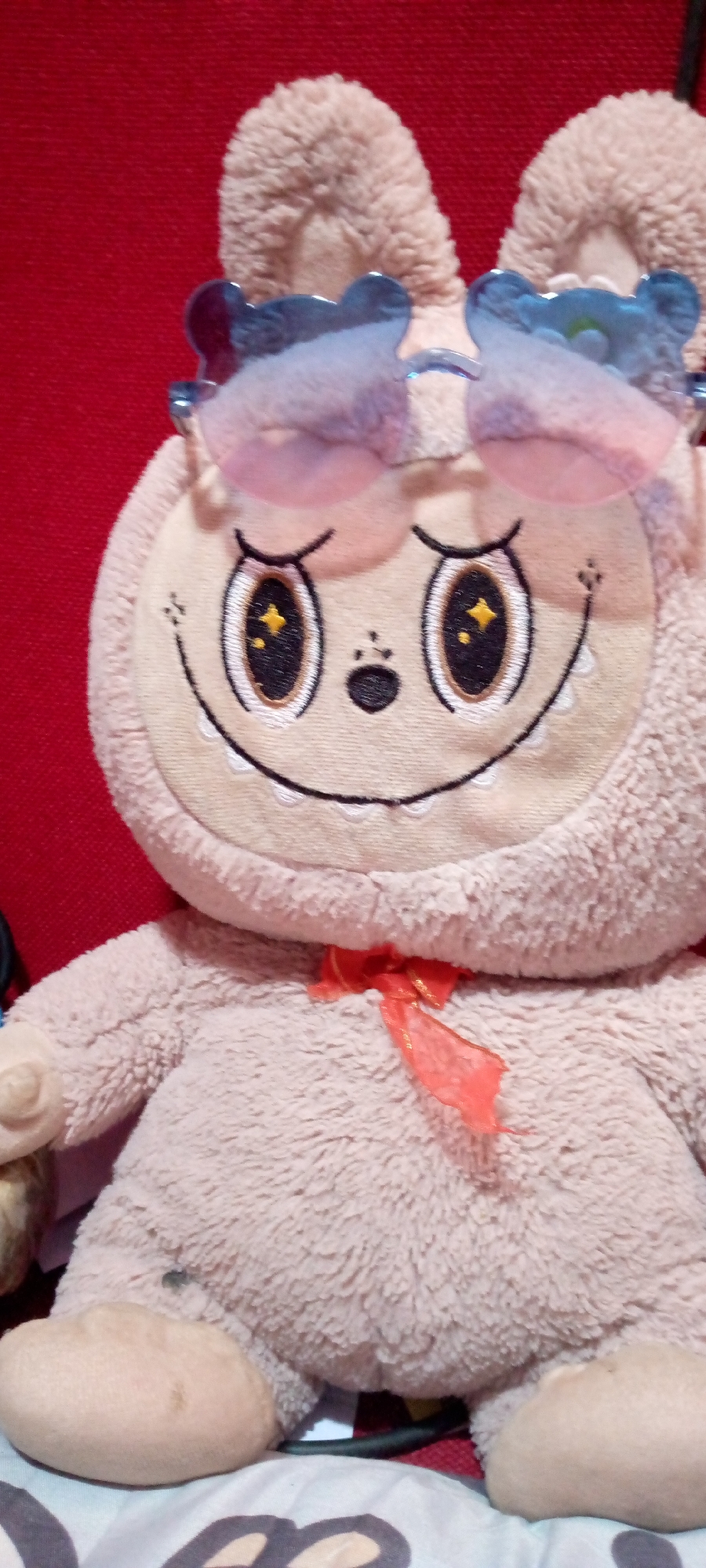 A close-up, eye-level, vertical, medium shot captures a light brown plush toy. The toy, resembling a rabbit or bear, is adorned with large, blue, translucent sunglasses with a heart-shaped outline and a wavy top edge. It wears a bright orange, sheer ribbon tied into a bow around its neck. The toy's face features embroidered details: closed eyes with yellow star-shaped pupils against a black background, small dots for cheeks, a small black nose, and a wide, black stitched smile with white scalloped teeth. The toy is positioned against a deep red background. The lighting appears to be artificial and casts soft shadows. No geographical context of Sumberwono, Indonesia, is evident in the visual information provided.
