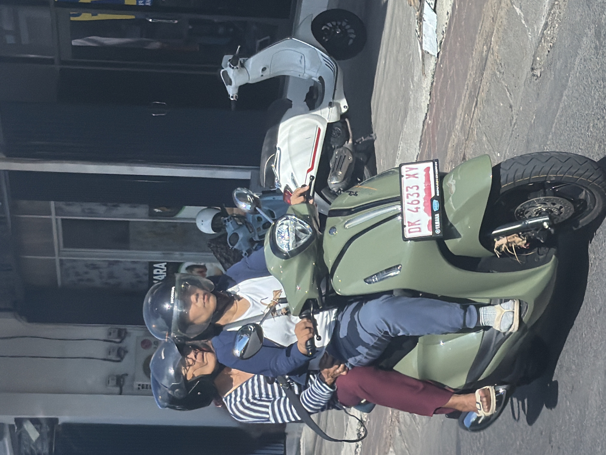 Two individuals, both wearing helmets, are seated on a green scooter, likely stopped on a street. The person in front, presumably the driver, wears a white t-shirt layered under a blue long-sleeved shirt and blue pants, with light-colored sneakers. The passenger behind them wears a horizontal striped long-sleeved shirt and maroon pants, with sandals on their feet.
The scooter they are on is an olive green color, and its license plate, visible on the rear fender, reads "DK 4633 XY". The license plate frame also displays "YAMAHA" and the motto "SEMAKIN DI DEPAN". Another white scooter, resembling a Vespa, is parked behind them, and a blue scooter is partially visible further back.
The scene appears to be set on a paved street or sidewalk, alongside buildings with dark windows. Strong sunlight indicates it is daytime, casting clear shadows. A "HONDA" logo is visible on the side of one of the helmets. Partially visible text on a sign in the background reads "ARA". The license plate code "DK" suggests a location in Bali, Indonesia.