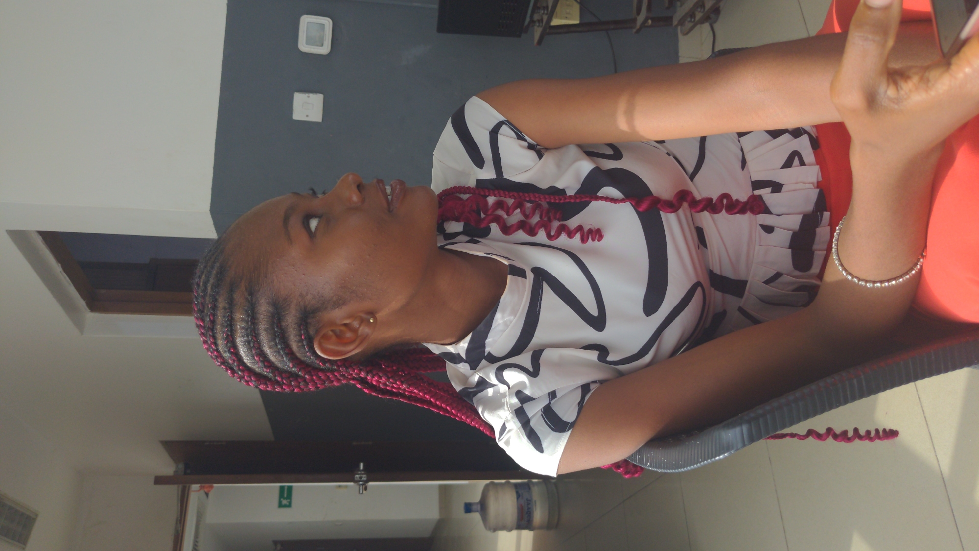 A young person with dark skin is depicted in a close-up, vertical shot, looking upwards and slightly to the left with an open-mouthed expression, suggesting they are speaking or reacting. The setting is indoors, likely in Abuja, Nigeria, and well-lit, indicating daytime.
The subject has prominent pinkish-red braided hair, styled in neat cornrows at the front that transition into loose, spiral curls. They are wearing a white short-sleeved top with a bold, abstract black pattern and a pleated detail on the side. A silver-toned bracelet with small beads adorns their right wrist, and a subtle gold stud earring is visible in their left ear. Their right hand is partially visible, appearing to hold a mobile phone.
The background features a two-toned wall: light grey on the upper section and a darker grey or charcoal on the lower half. A small, dark-paned window is set high on the light grey wall. On the darker wall, two white square devices, possibly light switches or sensors, are mounted. A green exit sign symbol (a white running figure) can be seen on the lower part of the dark wall. On the floor, a large water dispenser with a plastic bottle, partially showing a "LAQUA" brand label, is visible. The person is seated in a dark-colored chair.