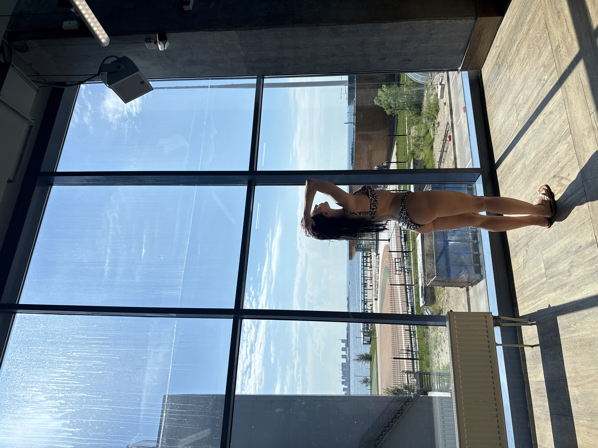 A woman in a leopard-print bikini stands with her hands behind her head, looking out a large window at a body of water with a city skyline in the distance. The window overlooks an outdoor area with a running track and some industrial elements. The scene appears to be within a modern building with concrete walls and a wooden floor. The natural light filtering through the window casts shadows across the floor. The window glass is dirty with streaks. The city visible in the distance is Saint Petersburg, Russia.