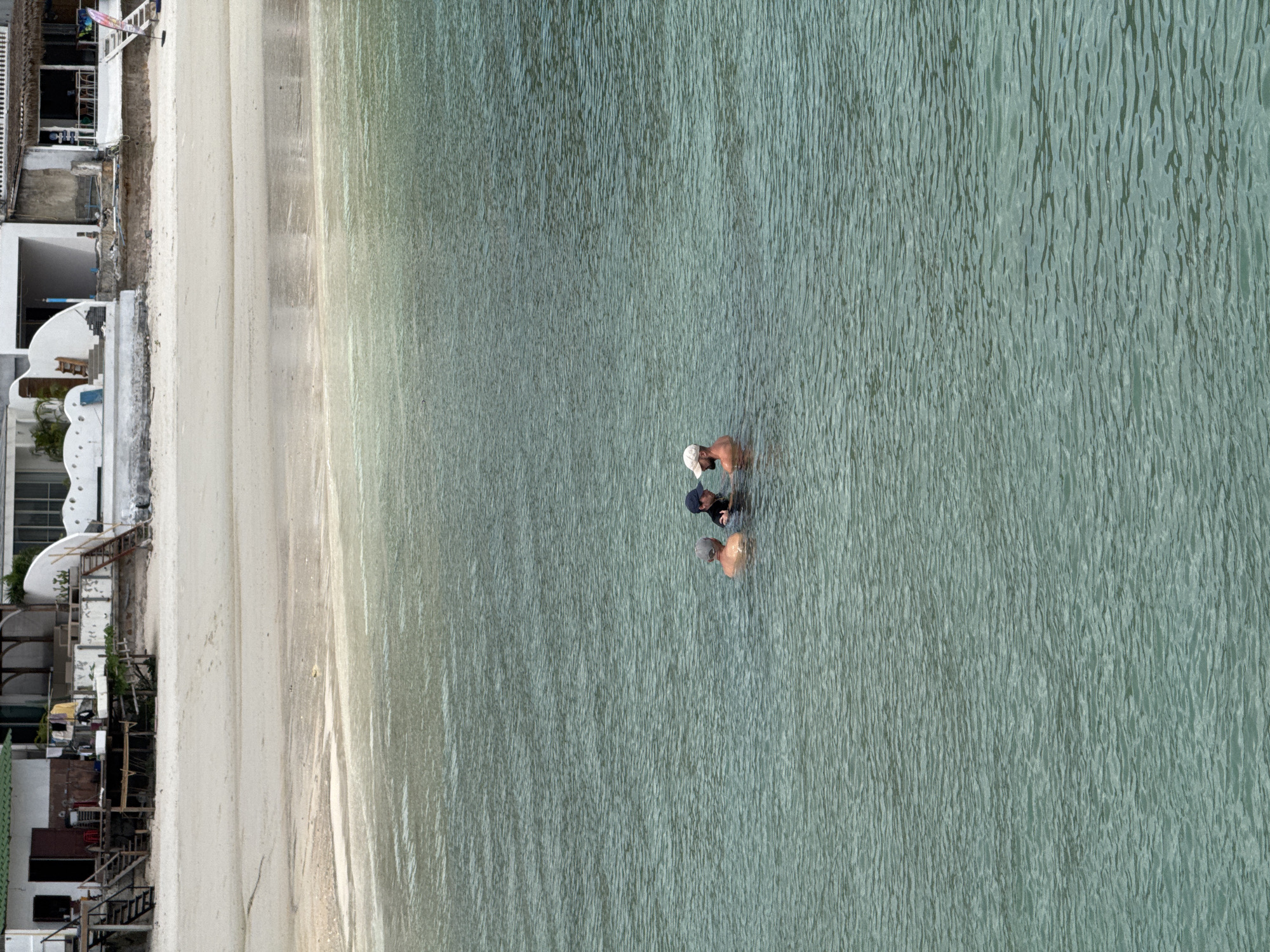 In the waters off the coast of ตำบลเกาะพะงัน, Thailand, three individuals are partially submerged. Two are wearing baseball caps, one dark blue and the other a light beige. The third person is wearing a light grey cap. Their torsos and heads are visible above the surface of the clear, greenish water. In the background, a sandy beach is visible, backed by a row of buildings, some with green roofs and wooden structures, and others with white walls and stairs leading down to the beach. The scene suggests a relaxed atmosphere, possibly a recreational area.