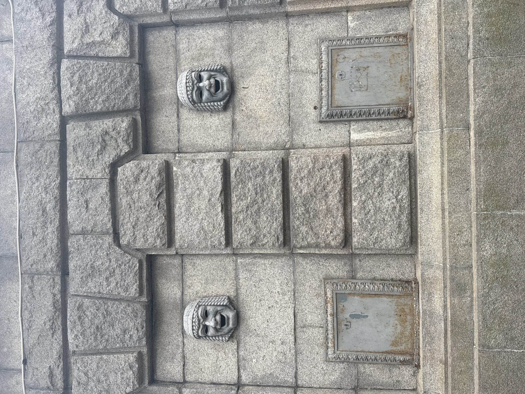 The image captures a detailed section of a weathered grey stone wall, likely granite, showcasing architectural ornamentation. The wall is constructed from large, roughly cut blocks, interspersed with smoother, recessed sections that create a layered, stepped pattern.
Two identical sculpted faces, resembling ancient theatrical masks, project from the wall. Positioned vertically one above the other, each mask depicts an open-mouthed, seemingly crying or distressed expression, framed by stylized hair or headwear. These decorative elements are rendered in a slightly lighter grey material than the surrounding stone.
To the right of the masks are two rectangular metal plates, also arranged vertically. Each plate features an ornate, decorative border surrounding a plain, flat central panel. Both central panels show signs of age, with some discoloration and minor rust, and each includes a small keyhole on its left side. These plates likely serve as access covers for utilities.
A thin, light-colored cable runs vertically along the far left edge of the wall. The lighting is soft and even, suggesting either an overcast day or a shaded area, without harsh shadows. The robust granite construction and classical-inspired masks and ornate metalwork are consistent with historical architecture found in Saint Petersburg, Russia. There is no visible text in the image.