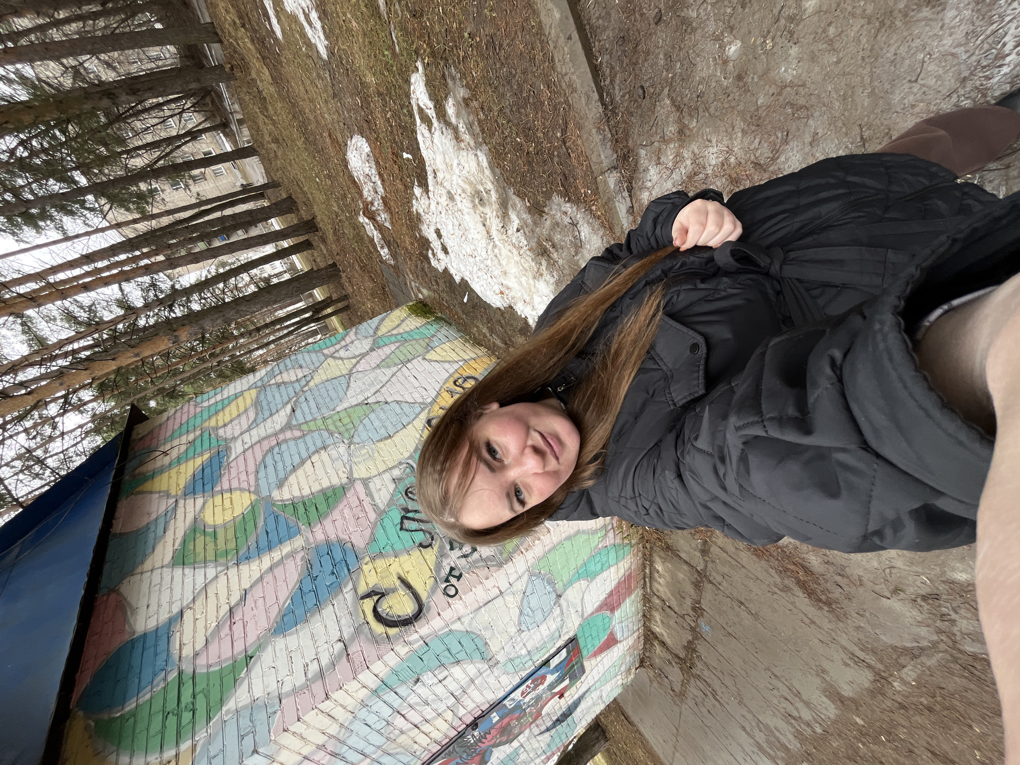 A woman with long brown hair, wearing a black jacket, is standing near a wall with a colorful mosaic design. The mosaic consists of numerous small, differently colored tiles arranged in a pattern resembling feathers or leaves. Some of the tiles are painted with images, including graffiti. The area around the woman and the mosaic has a mixture of grass, leaves, and patches of snow, suggesting it is late winter or early spring. In the background, tall bare trees and a building are visible. The location is the city of Chaikovsky, Russia.