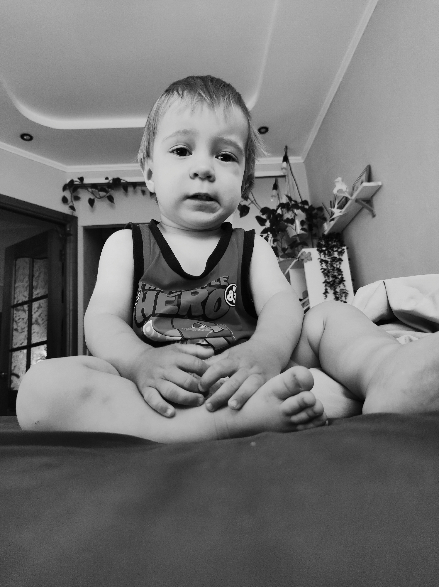 A toddler is sitting on a bed. The location is likely in the city of Chaikovsky, Russia. The child is wearing a tank top with the word "HERO" printed on it. They have fair skin and short blonde hair. The child is looking directly at the camera with a slightly pensive expression. The background is a room with a door on the left and plants on a shelf on the right. The image is in black and white.