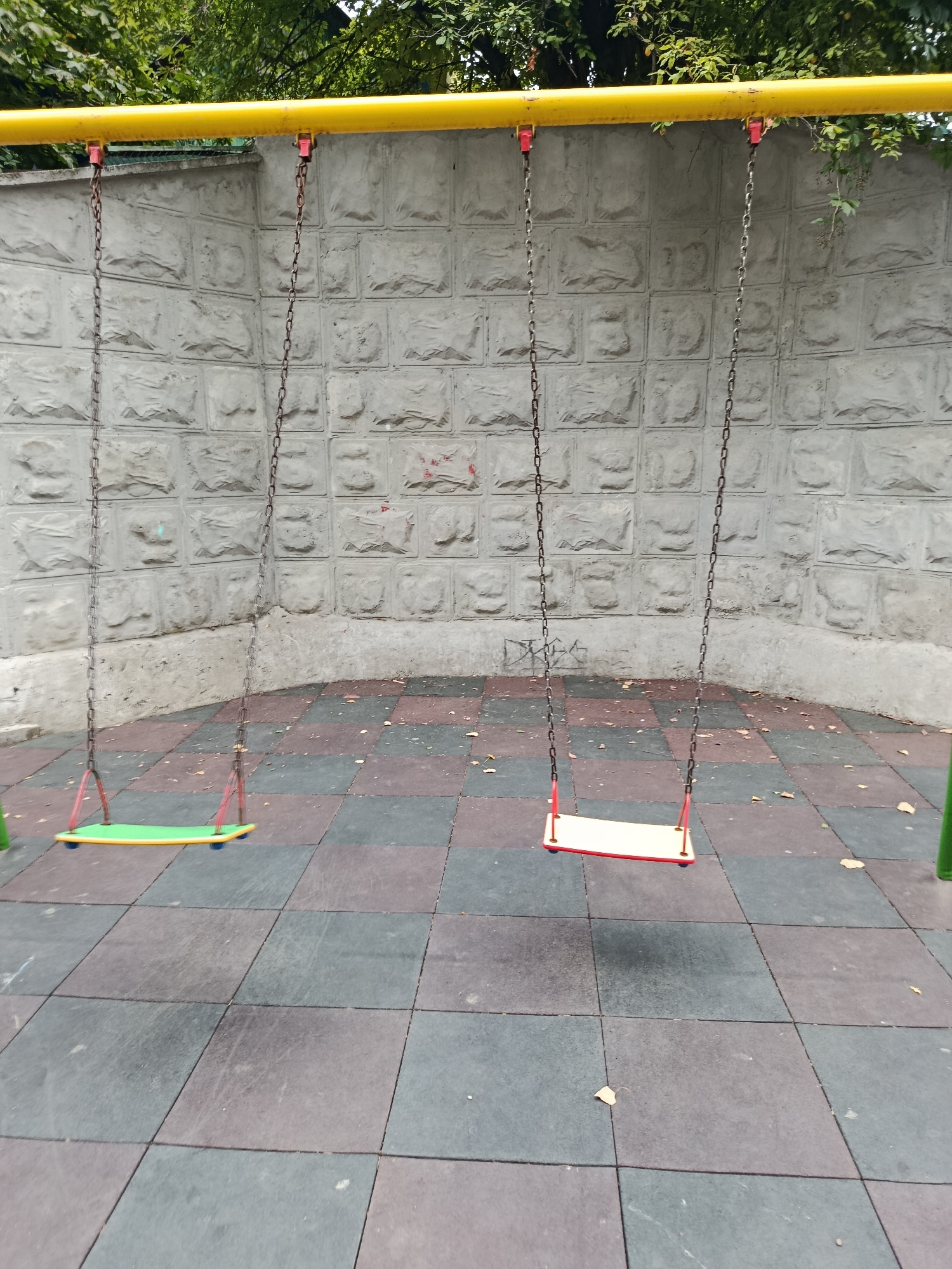 A public playground in Chișinău, Moldova, features a yellow metal swing set with three swings hanging from metal chains attached to the yellow bar. The swings have red metal frames and seats; one is green and the other two are yellow and red. The ground beneath the swings is a patchwork of grey and black rubber safety tiles. Behind the swing set is a textured concrete wall, and above that, green foliage from trees is visible. The scene appears to be in a state of disuse, with a few scattered leaves on the ground.