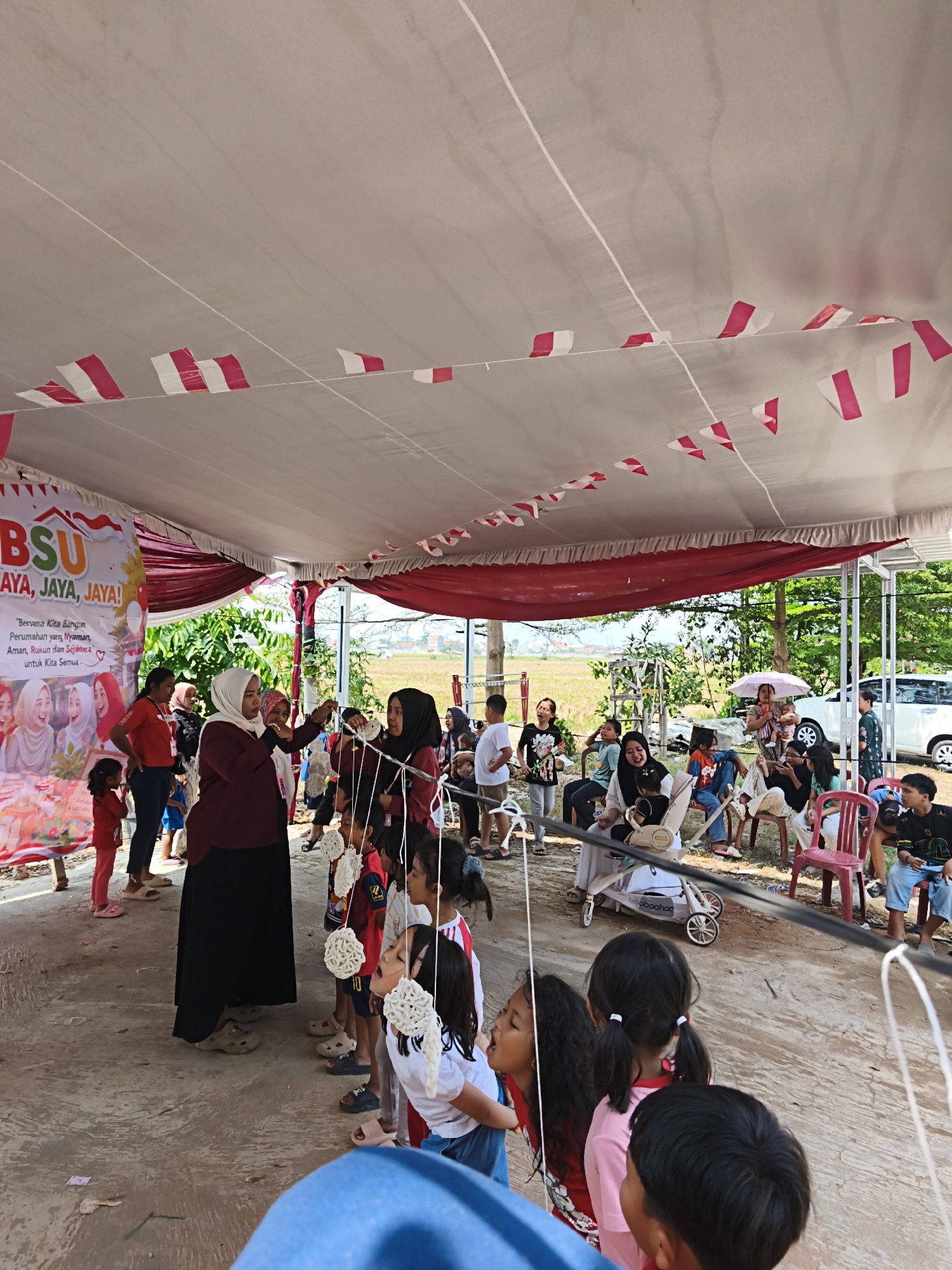 The image depicts an outdoor event, possibly a festival or competition, held under a large white tent decorated with alternating red and white pennant flags. In the foreground, a group of children are participating in a game, their faces focused on something happening off-camera. Several adults are also present, some observing the children and others appearing to be involved in organizing or supervising the event.
A banner in the background, partially visible, displays the text "BSU JAYA, JAYA, JAYA!" along with images of people. The ground is paved with concrete. Beyond the tent, a field of dry grass is visible, suggesting a rural or semi-rural setting. Several vehicles, including a white car, are parked in the background, and a few buildings can be seen in the distance. The overall atmosphere appears festive and community-oriented.
While the image itself does not explicitly state its location, if this event is indeed in Bandar Lampung, Indonesia, it would be an outdoor public space or community gathering area within the city. The presence of the red and white pennant flags could indicate a celebration of Indonesian Independence Day or another national event.