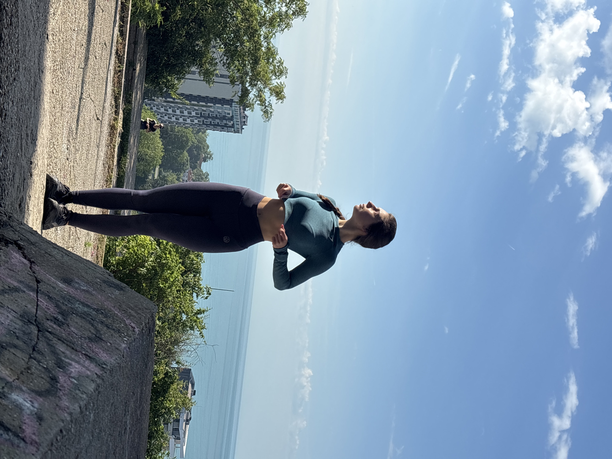 A full-length shot depicts a young woman standing outdoors in Varna, Bulgaria, on what appears to be a bright, sunny day. She is dressed in athletic wear, consisting of a dark teal long-sleeved crop top and dark leggings with matching sneakers. Her dark hair is visible, possibly in a braid, over her shoulder. The woman stands with her hands near her waist, looking upwards and slightly to her right, with a calm and contemplative expression.
She is positioned on a paved pathway or concrete platform, with a rough concrete barrier or wall to her left. In the background, a vast, calm body of blue water stretches out, likely the Black Sea, meeting a distant horizon. Along the coastline in the distance, modern-looking high-rise buildings are visible amidst green trees and foliage. The sky above is clear and bright blue, dotted with scattered white clouds. Another, smaller figure is faintly discernible further down the paved area in the mid-ground. The overall scene conveys a sense of peaceful outdoor activity or enjoying a scenic coastal view. There is no visible text in the image.