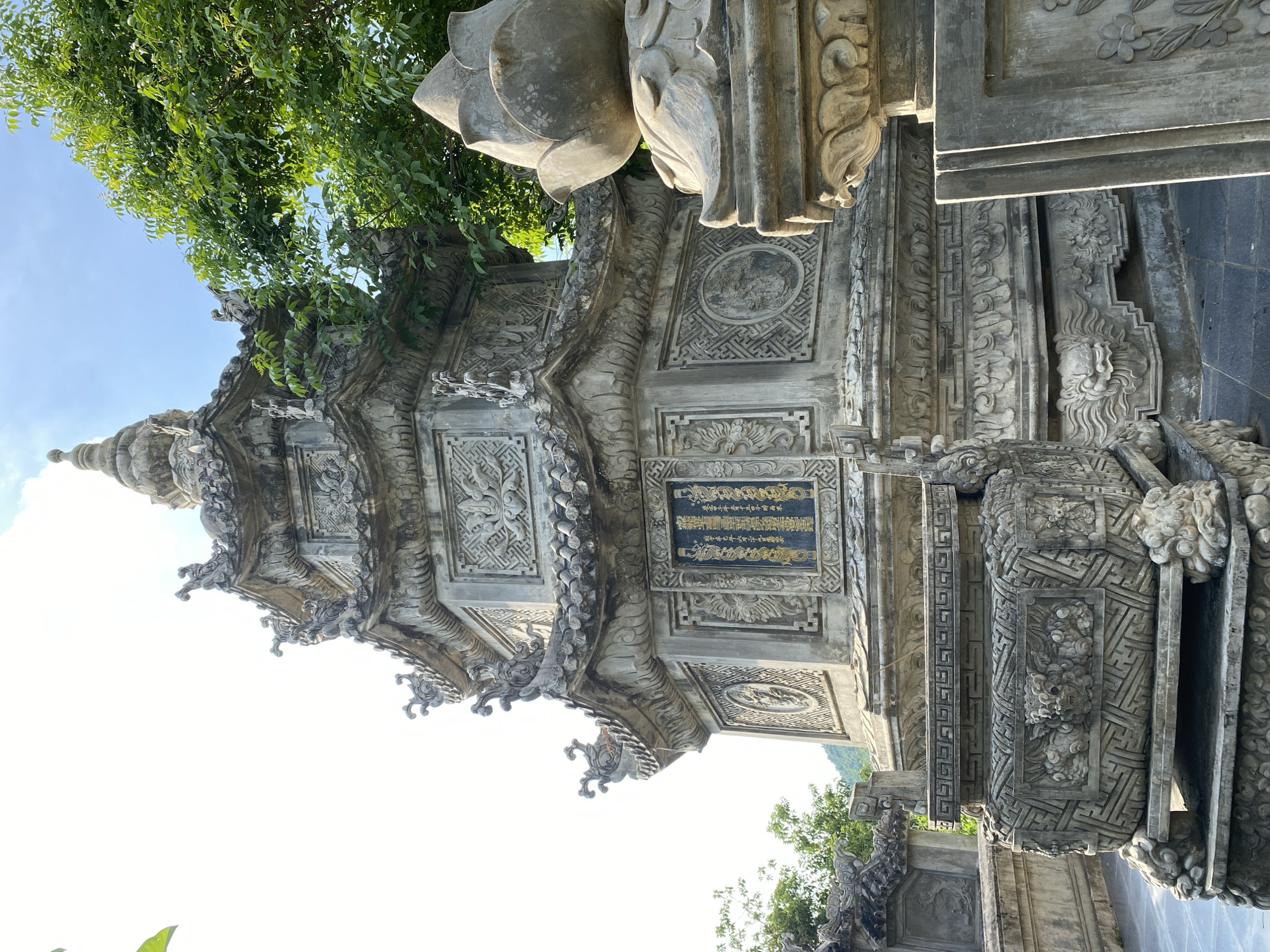 The image depicts a stone stupa located in Tay Nha Trang Ward, Vietnam. The stupa is intricately carved with ornate details, including mythological creatures, floral motifs, and geometric patterns. A black plaque with golden inscriptions is visible on one of the stupa's faces. In the foreground, a stone incense burner with similar decorative carvings rests on a tiered base. To the right, a sculpted lotus flower on a pedestal is partially visible. Lush green foliage from nearby trees overhangs the scene, and a clear blue sky with sparse clouds is visible in the background. The ground is paved with dark square tiles. The overall scene suggests a peaceful, spiritual, or historical site, likely a temple or pagoda.