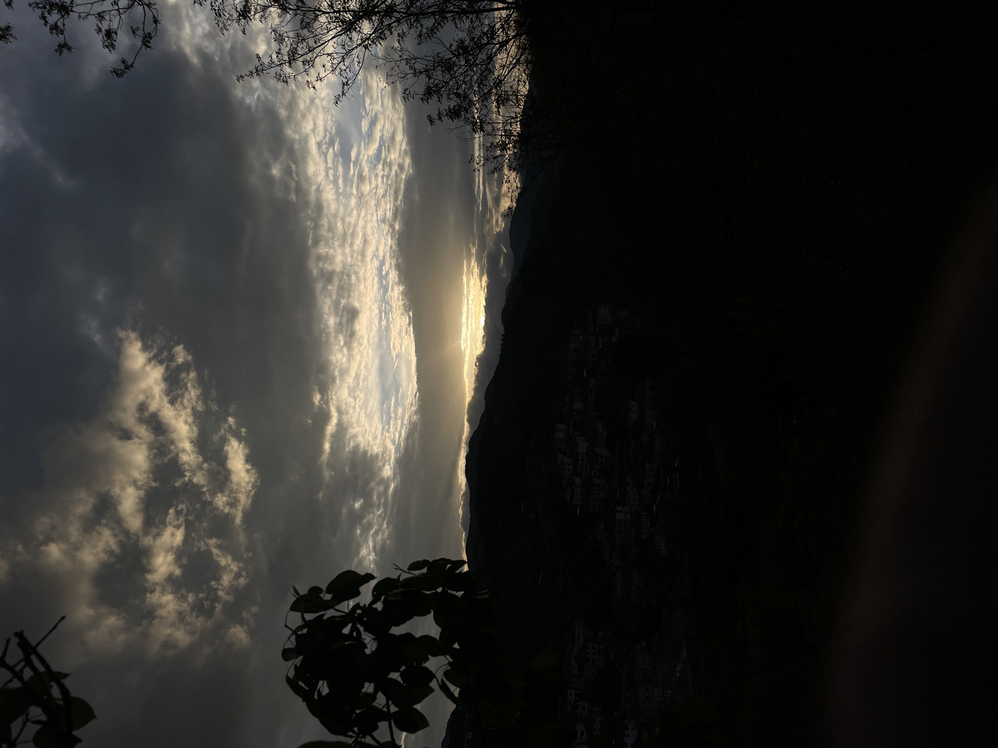 The image shows a view towards a mountain from a low-angle perspective, likely taken near Zlatograd, Bulgaria. The sky occupies a large portion of the frame, displaying a mix of dark, ominous clouds and patches of bright sunlight. The light is most prominent along a horizon line formed by the mountain silhouette. The mountain itself appears mostly dark, its details obscured by the setting sun, although some buildings are visible lower down on the mountainside. Silhouetted trees and foliage are visible at the top and bottom corners of the image. The overall scene is dark and dramatic.