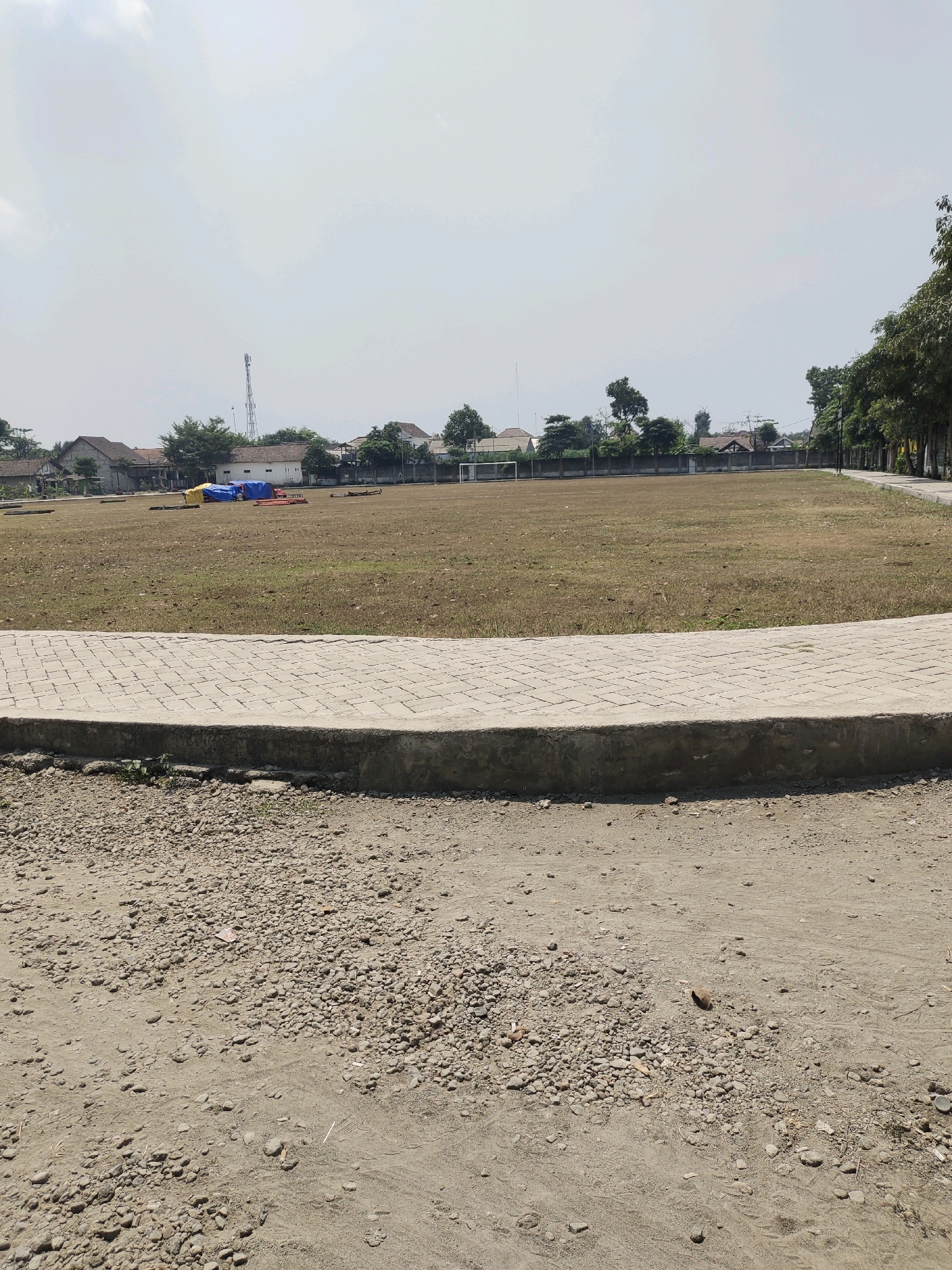 The image depicts an outdoor scene, likely a sports field or park area, located in Ringinrejo, Indonesia. In the foreground, a paved walkway with a curved edge is visible, constructed from light-colored rectangular bricks. The walkway sits on a concrete base. Below the walkway, the ground is covered in loose gravel and dirt, with some small patches of dry grass.
Beyond the walkway, a large, dry, brown field stretches into the midground. Scattered across this field are several objects, including what appear to be tarpaulins in blue and yellow, as well as some metallic structures. In the distance, a fence delineates the edge of the field. Behind the fence, a row of buildings, including houses with tiled roofs and a white building with a tower, are visible. Trees are interspersed among the buildings and along the right side of the image, lining a paved pathway or road that curves away into the distance. The sky is bright and overcast, suggesting a daytime scene. No people are visible in the image. The environment appears to be a community or public space in a somewhat rural or suburban setting.