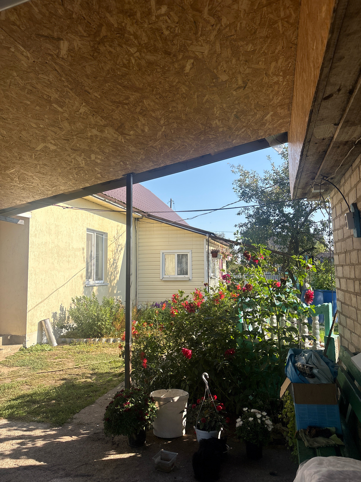 The image depicts an outdoor scene in the area of Хорастаўскі сельскі Савет, Belarus. In the foreground, a gray concrete surface is partially shaded by an overhead structure with a wood-chip covered ceiling. A white bucket and a black potted plant are visible on the concrete. To the right, a brick wall forms the side of a building, with a green wooden bench and some stacked boxes and materials. Beyond this, a dense area of flowering bushes with red and pink blooms is present. In the middle ground, a yellow house with a brown roof is visible, featuring white-framed windows. Behind the house, a large tree with green foliage is prominent against a clear blue sky. The overall impression is that of a backyard or garden area adjacent to a residential structure.