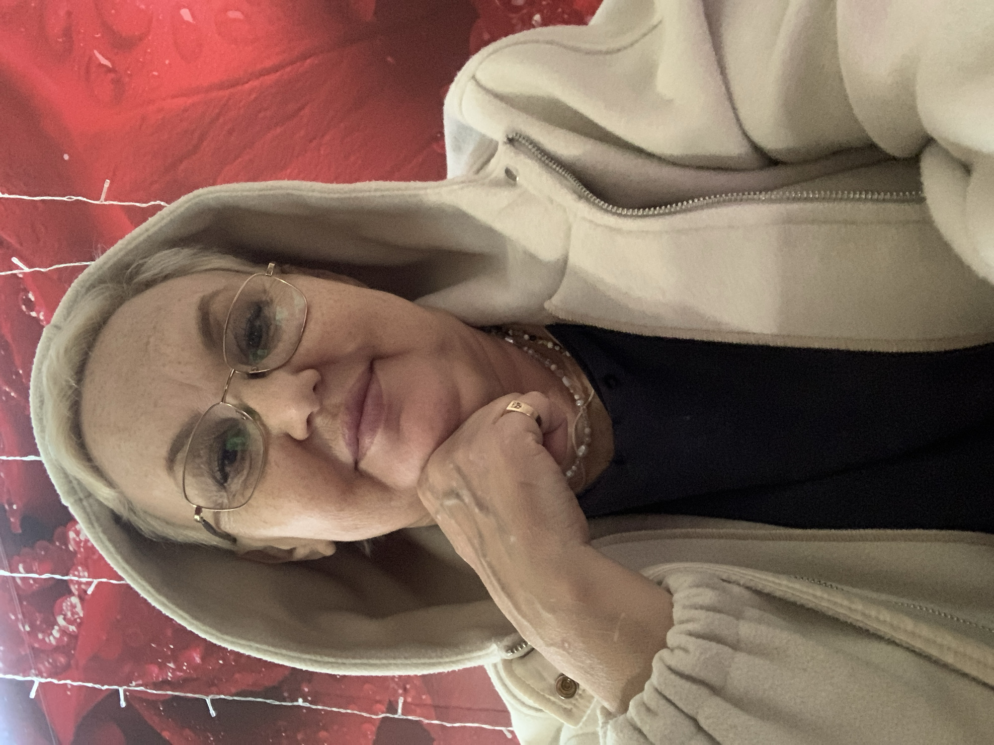 A woman with fair skin and blonde hair is wearing glasses and a hooded sweatshirt. She is resting her chin on her hand, which has a gold ring on one of the fingers. She also has a pearl necklace on. The background is red with white lights and what appears to be a rose pattern. The location appears to be Chaikovsky, Russia based on previous instructions.