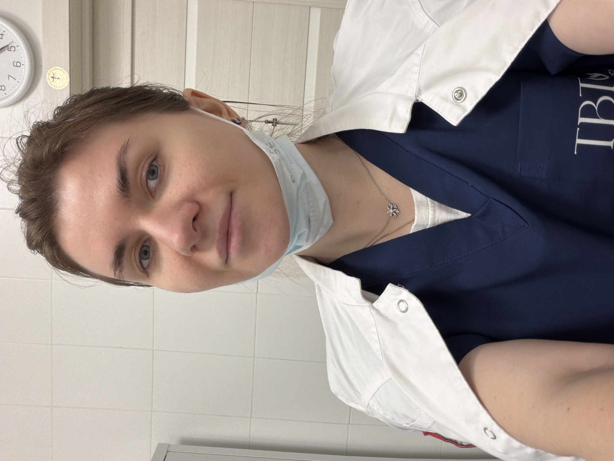 A young woman, likely in her 20s or early 30s, is captured in a vertical, close-up selfie within what appears to be a professional indoor setting. She has fair skin, light brown hair pulled back, and blue-grey eyes, looking directly at the camera with a slight, neutral expression. She is wearing a white lab coat or similar professional jacket with snap buttons over a navy blue scrub top. A light blue surgical mask is pulled down and rests below her chin.
Around her neck, she wears a delicate silver chain with a snowflake-shaped pendant. A glimpse of an earlobe and a thin wire or hair accessory is visible behind her left ear. The navy blue scrub top features white text on the right side of her chest, partially visible as "TBLI" or "TBLY" followed by other letters, suggesting a logo or institutional name.
The background consists of white tiled walls, characteristic of a sterile or clinical environment such as a hospital, clinic, or laboratory. On the wall behind her, a white analog clock displays the time as approximately 12:45. Adjacent to the clock, a circular golden sticker with a cross-like or symbolic emblem is affixed to the wall. The bottom edge of the image shows part of a light-colored surface, possibly a counter or cabinet. The overall setting suggests a healthcare professional in a medical facility, consistent with a location like Khimki, Russia.