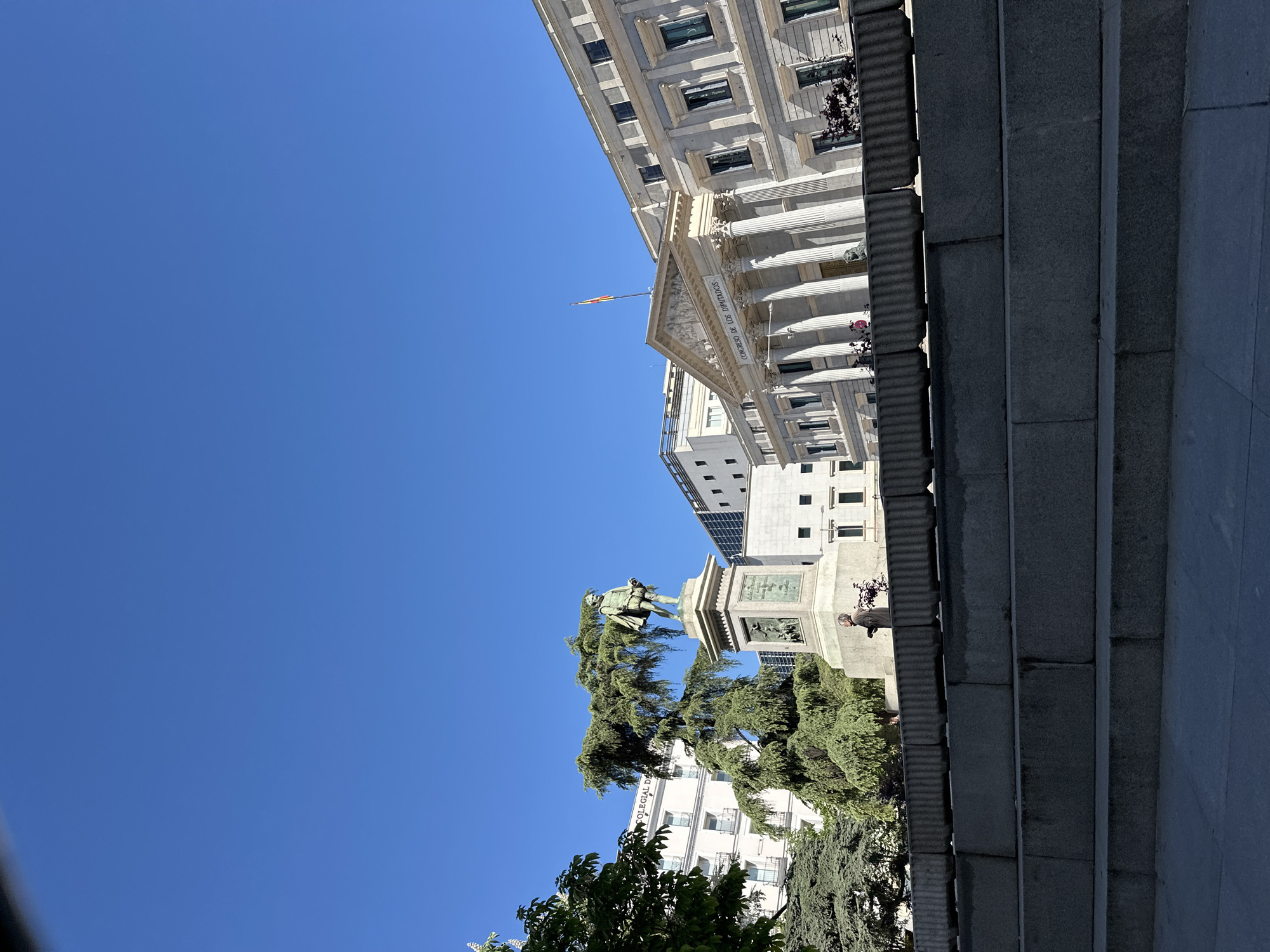 The image shows the Congress of Deputies in Madrid, Spain, viewed from below. The camera is pointed up and to the left. The building has a white facade with columns and a Spanish flag atop. A statue of Miguel de Cervantes stands in front of it, surrounded by green trees. Steps are visible to the right, leading up to the building. The sky is clear and blue.