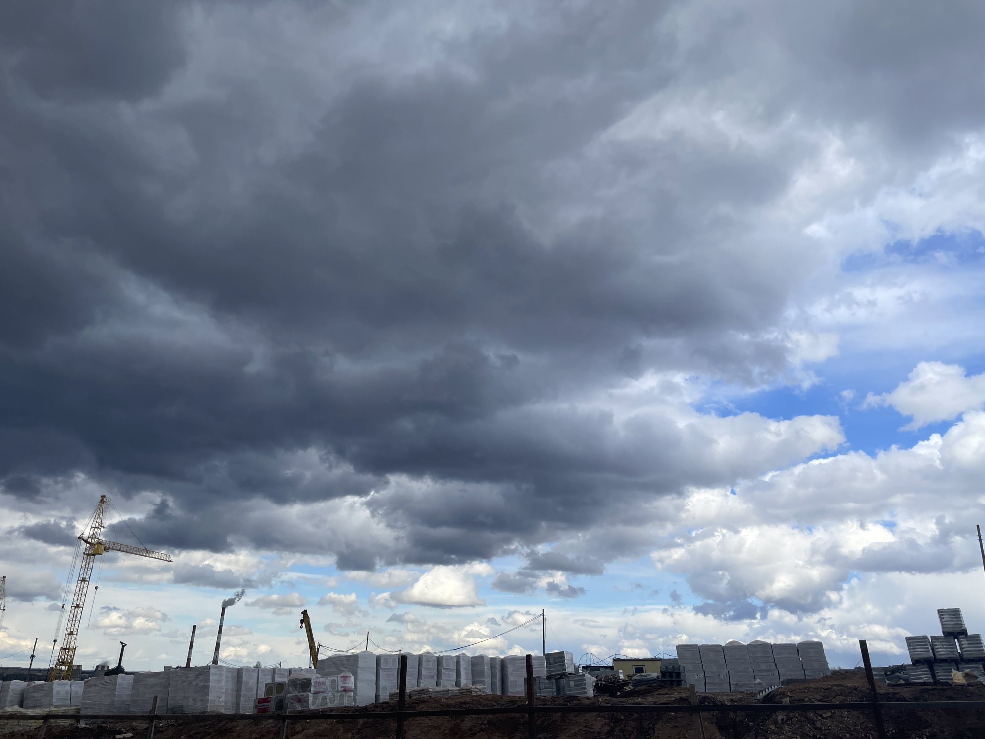 The image captures a construction site under a dramatic, heavily clouded sky in Pervomaysky, Russia. The upper and left portions of the sky are dominated by dense, dark grey storm clouds, while the right side transitions to lighter grey and white cumulus clouds with significant patches of bright blue sky peeking through. This contrast suggests changing weather, possibly before or after a rain shower, during daytime.
In the mid-ground, a large construction site stretches across the frame. Prominently on the left is a tall, yellow tower crane, its long arm extending horizontally. Further to the right, parts of another crane are visible. The ground is filled with numerous stacks of building materials, likely bricks or blocks, neatly arranged on pallets and wrapped in white protective plastic. Behind these materials, several tall, slender industrial chimneys or smokestacks rise into the sky, one of which is actively emitting a plume of dark smoke. Various smaller temporary structures and equipment are also scattered throughout the site. The foreground consists of rough, dark earth, possibly indicating recent digging, with some metal structures or fencing visible at the very bottom. No people are visible, and no discernible text is present in the image.