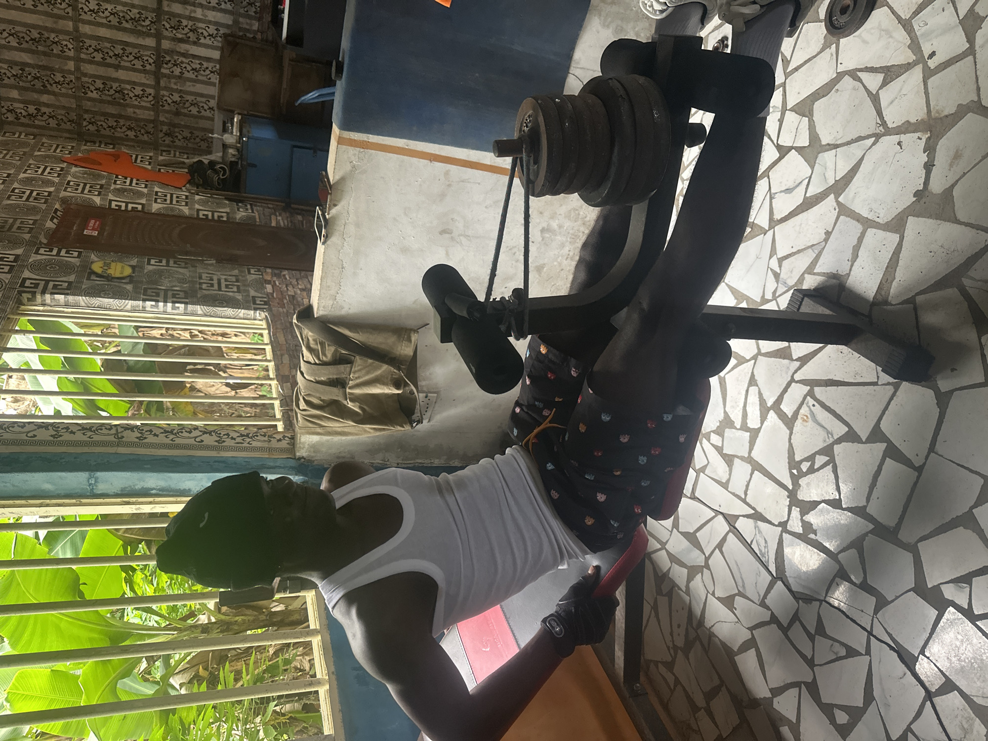 In an outdoor, covered space in Badagry, Nigeria, a Black male is seated on a weight training machine. He is wearing a white tank top, black beanie, and patterned shorts. He is wearing black gloves on his hands. The machine has a red padded seat and a black padded support for the legs. A black cable runs from the machine to a stack of weight plates. The floor is tiled with irregular white and grey pieces. Behind the man, a wall is decorated with patterned wallpaper and a blue cabinet is visible. Large green leaves from banana trees are seen through a barred opening. The lighting suggests it is daytime.