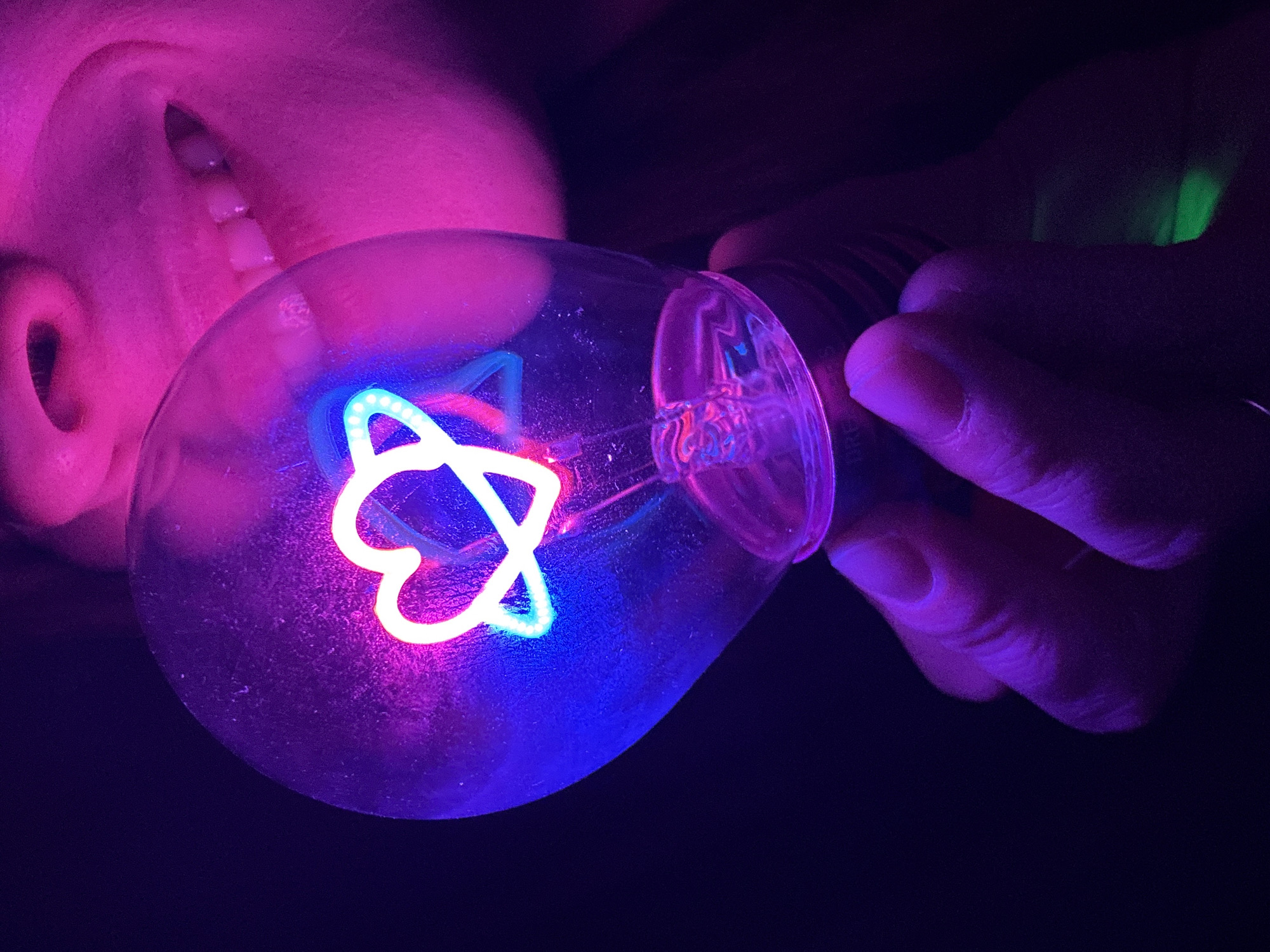 The image features a person holding a lightbulb in their hand. The lightbulb contains a neon sign that is shaped like a heart with an atom symbol in the middle. The person's face is visible in the background, with their mouth slightly open revealing teeth. The lightbulb is emitting a blue and purple glow, while the person's face is illuminated with a pink hue. There is a green light appearing on the hand holding the lightbulb. The background is dark. There is no indication of the city Budva, Montenegro.